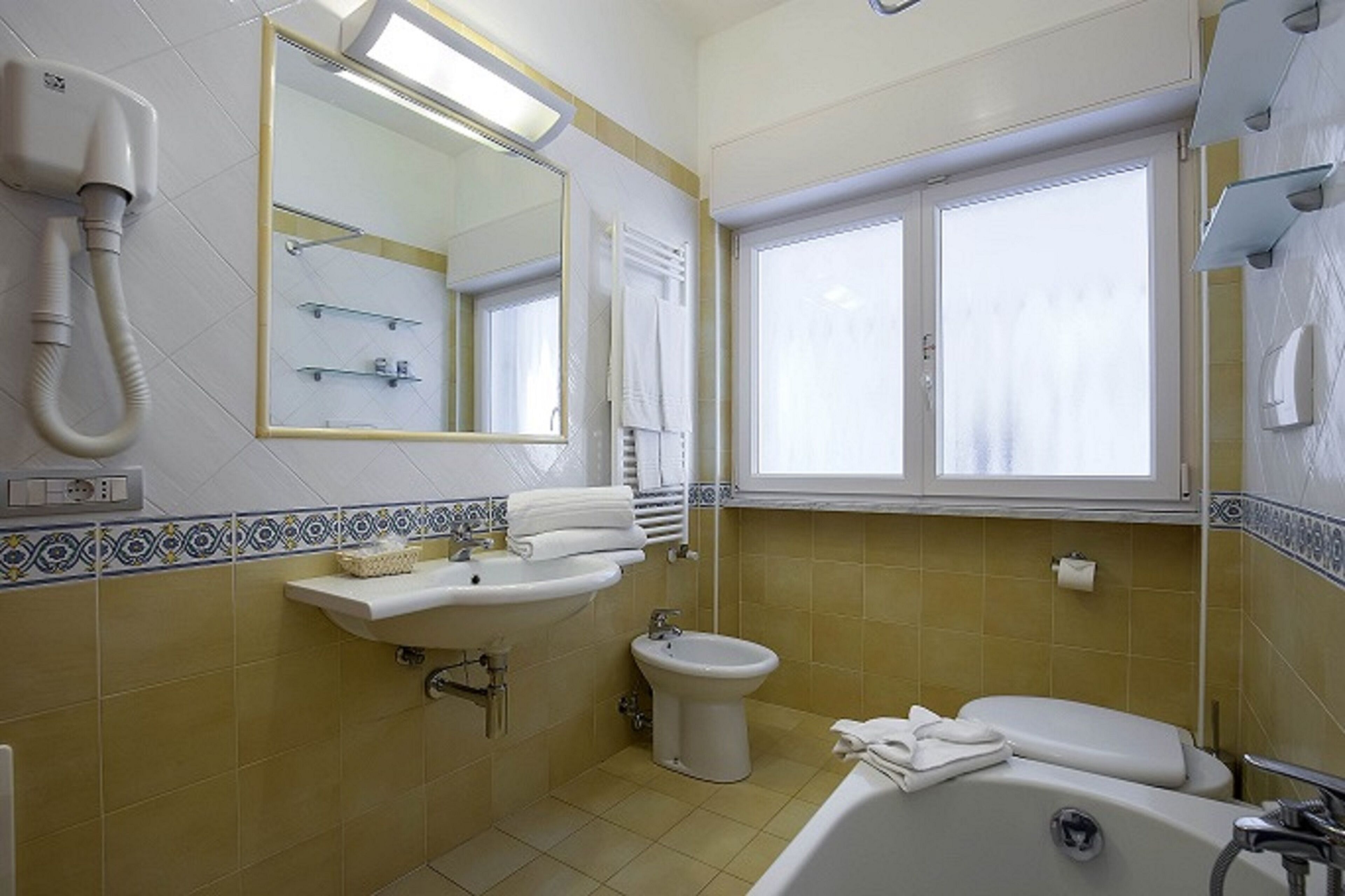 A bathroom with a sink, bidet, and bathtub. There is a window with curtains, a mirror, and a towel rack.