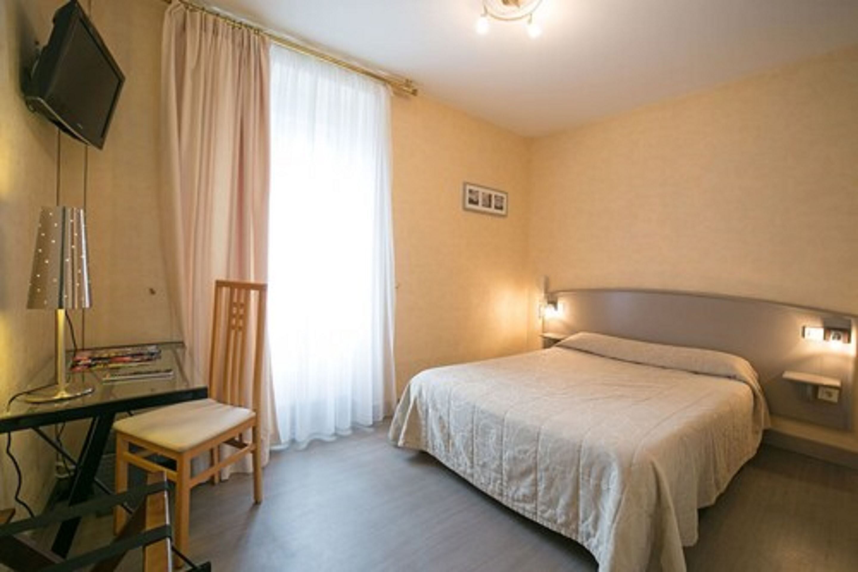 Una camera d'albergo con un letto, una scrivania con sedia, una televisione e una finestra con tende.