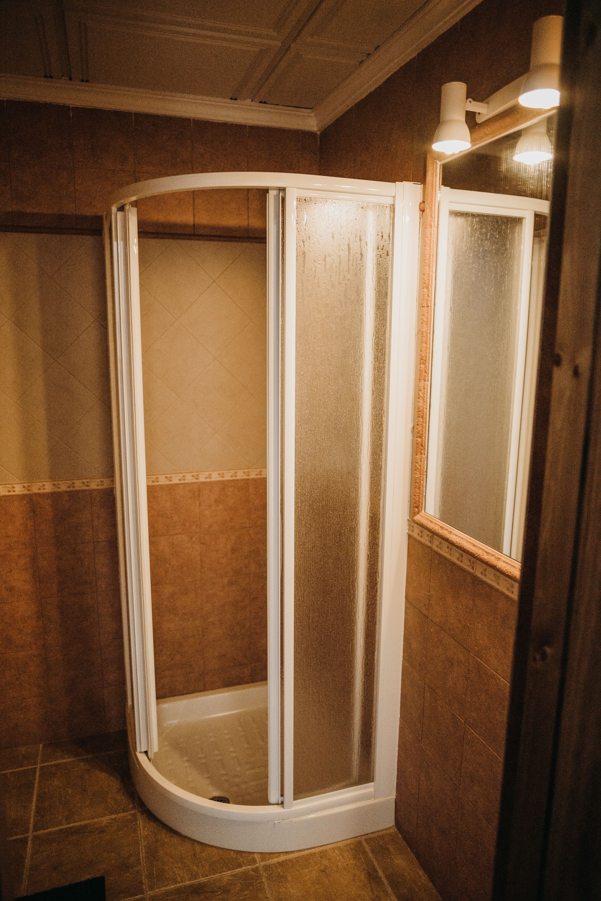 Ducha en esquina con puertas de vidrio esmerilado y marco de madera, ubicada en un cuarto con paredes de baldosas marrones y techo de madera.