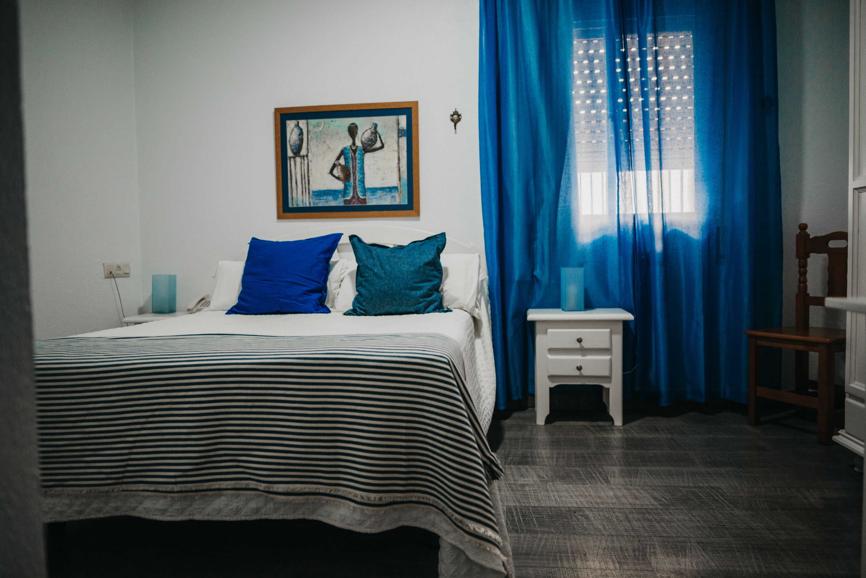 A bedroom with a bed, a nightstand, a chair, a painting on the wall, and blue curtains.