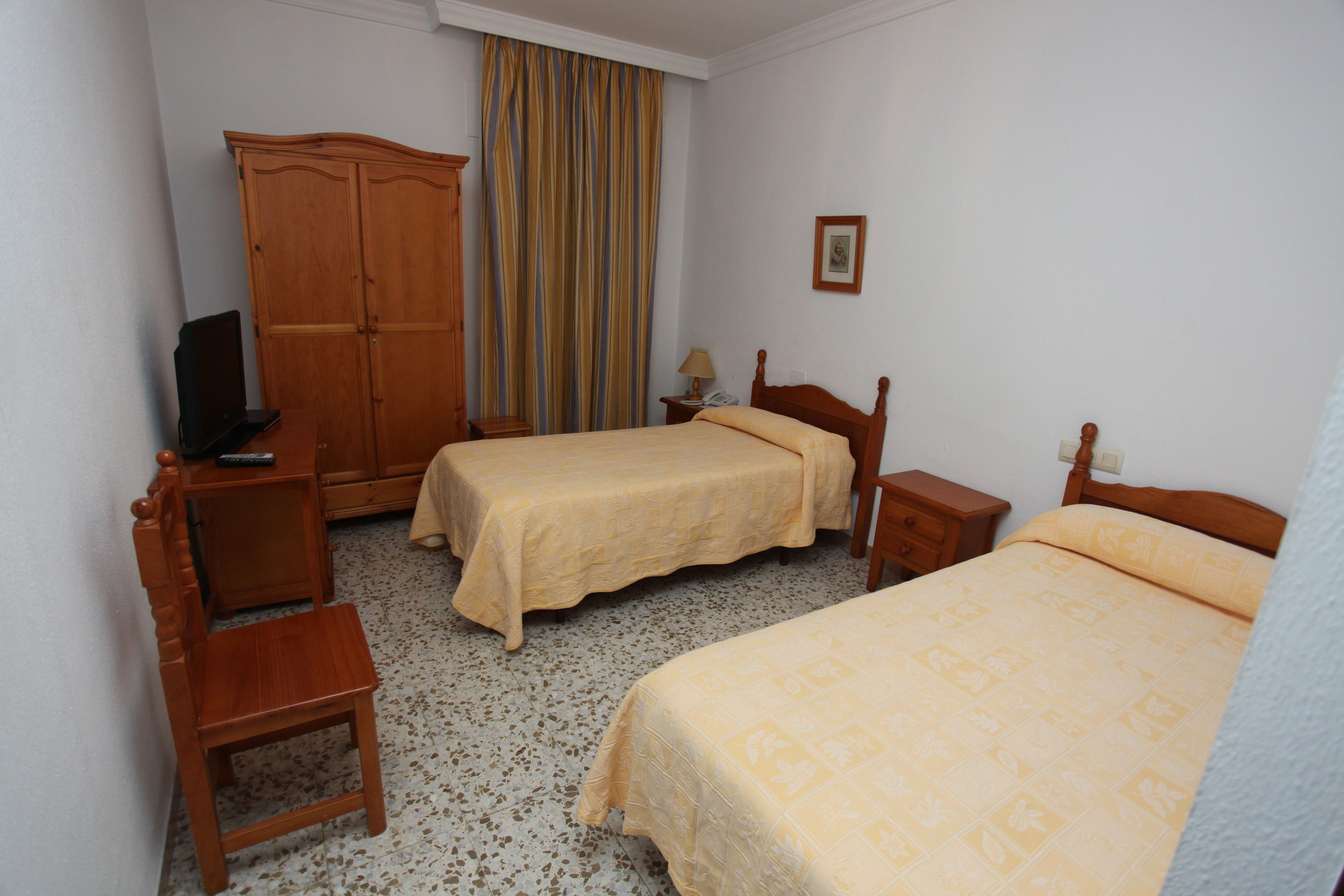 A hotel room with two single beds, a wooden wardrobe, a desk with a chair, a small table with a lamp, and a framed picture on the wall.
