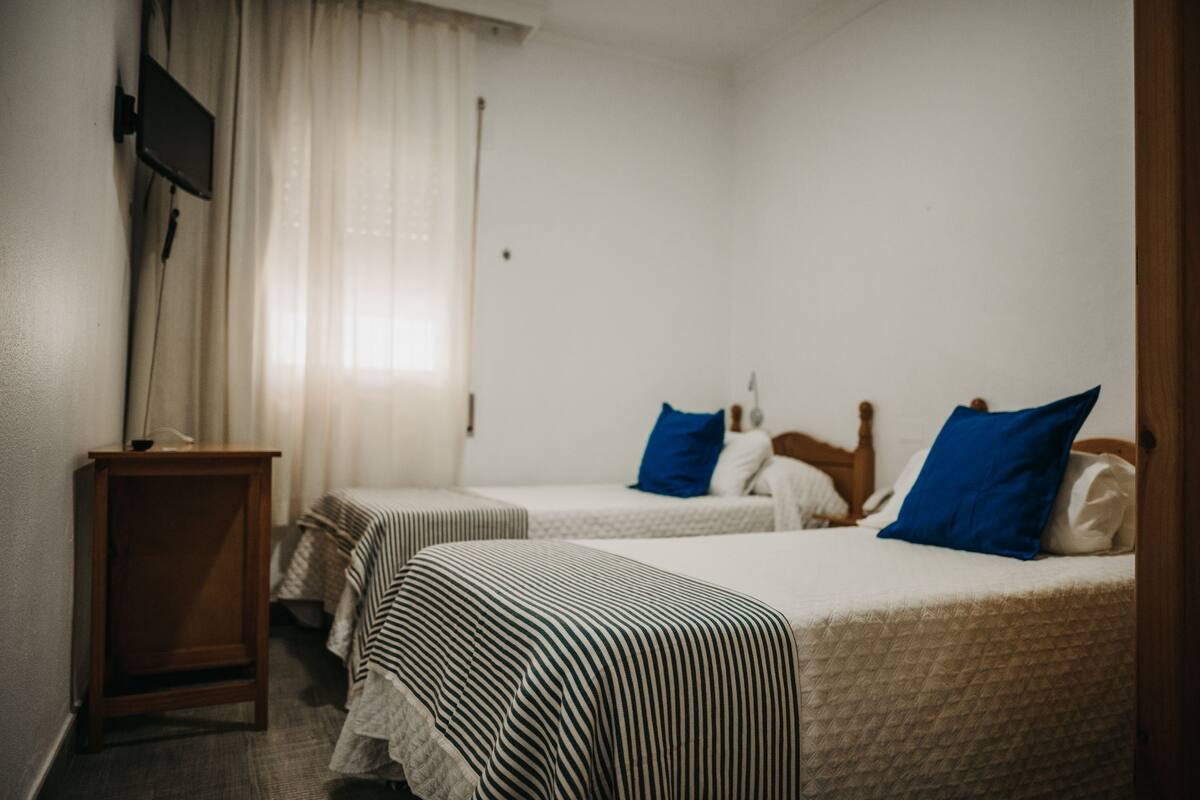 A room with two beds, a small wooden nightstand, a TV mounted on the wall, and a window with sheer curtains.