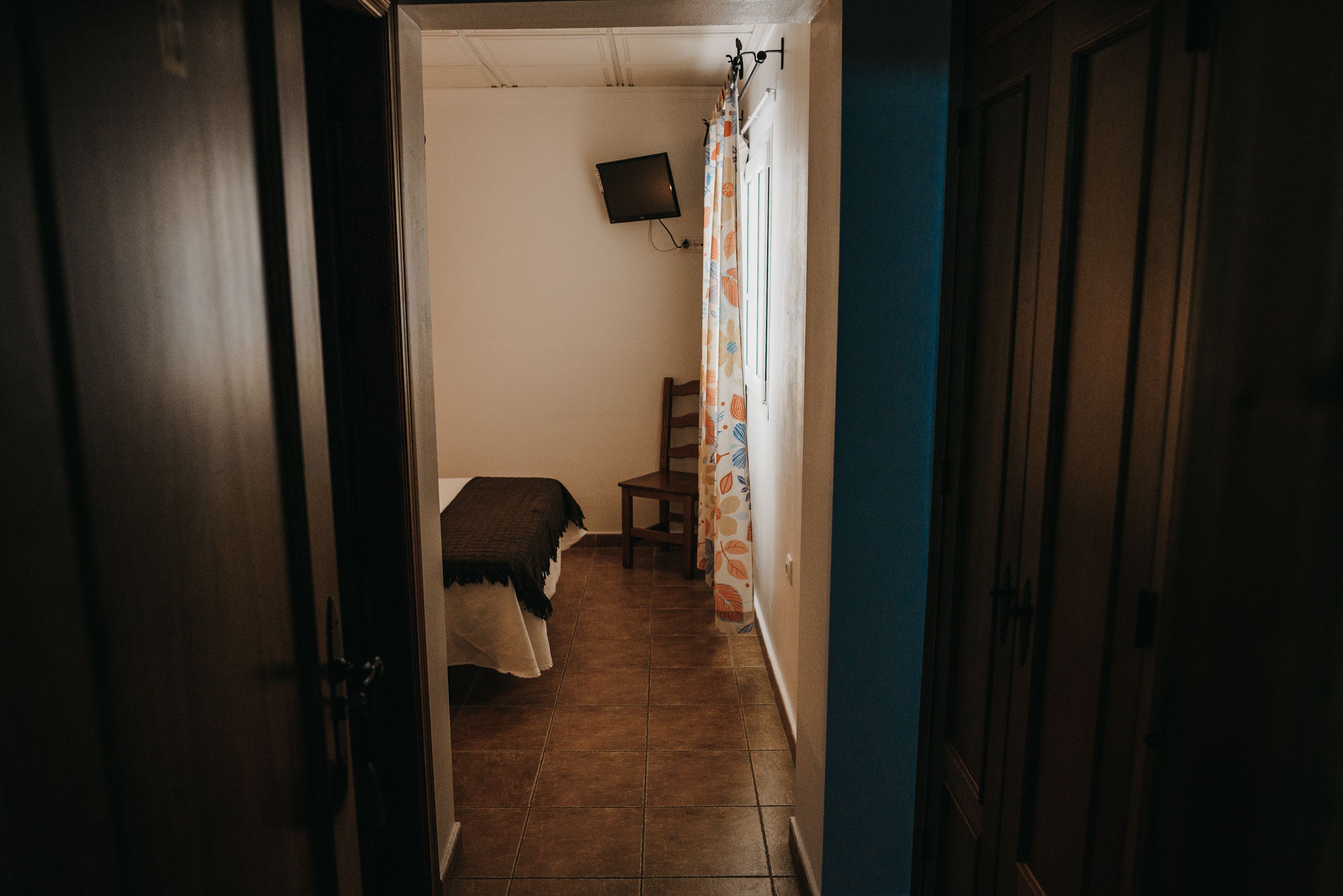 Un pasillo estrecho con una cama, una silla y un televisor instalado en la pared.