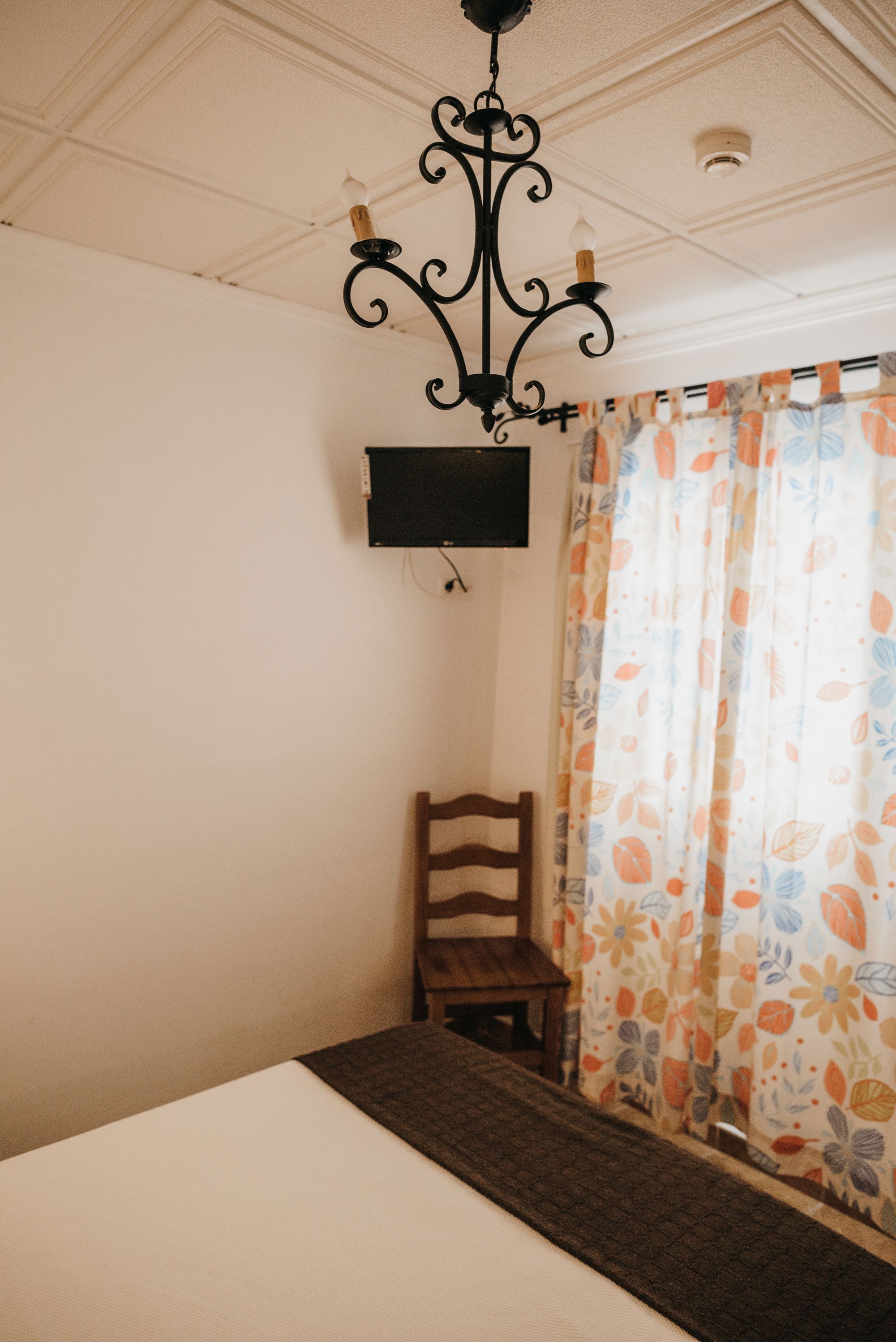 A room with a bed, a wooden chair, a television mounted on the wall, and a patterned curtain.
