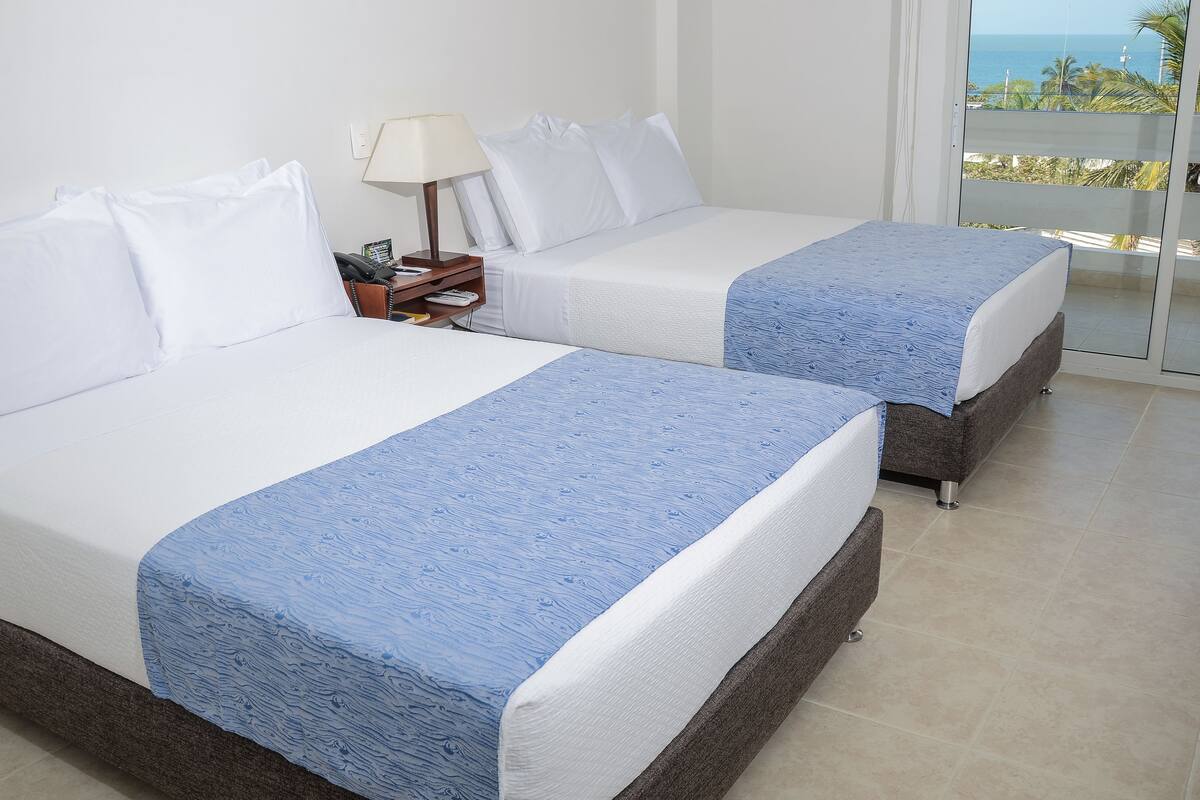 Dos camas con ropa de cama blanca y azul, una mesita de noche con una lámpara, y vista al océano a través de una puerta de balcón.