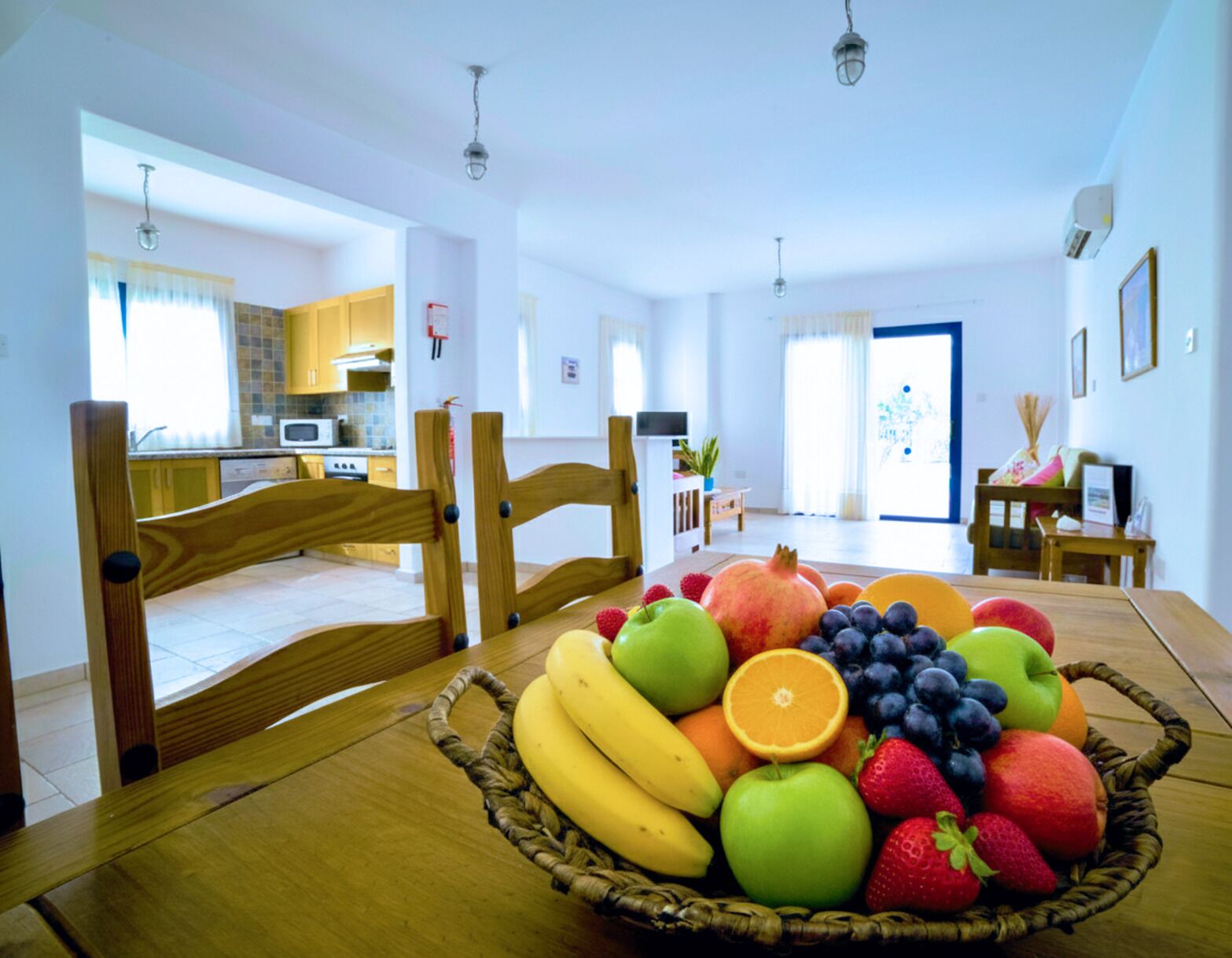 Un comedor con una mesa y sillas de madera, una cesta de frutas, y vistas a una cocina y salón.