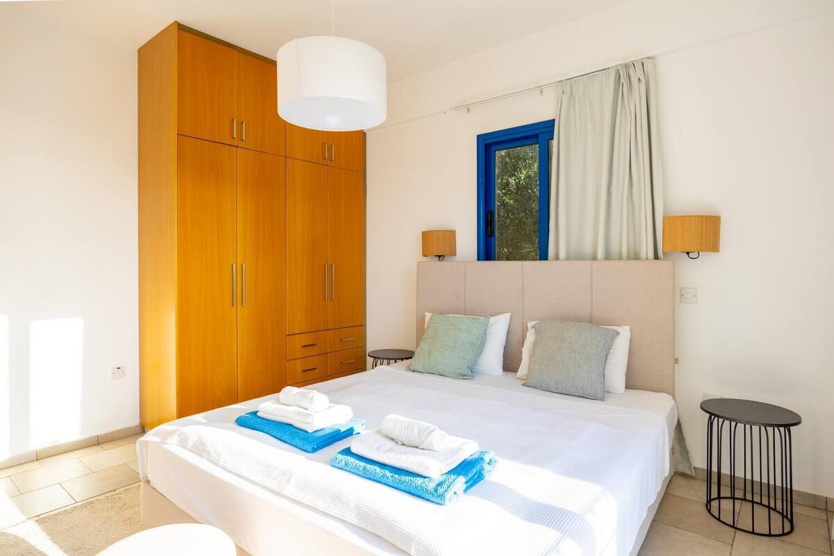 Een hotelkamer met een groot bed, een houten garderobe, twee bedlampen, een raam met gordijnen en een klein rond tafeltje.
