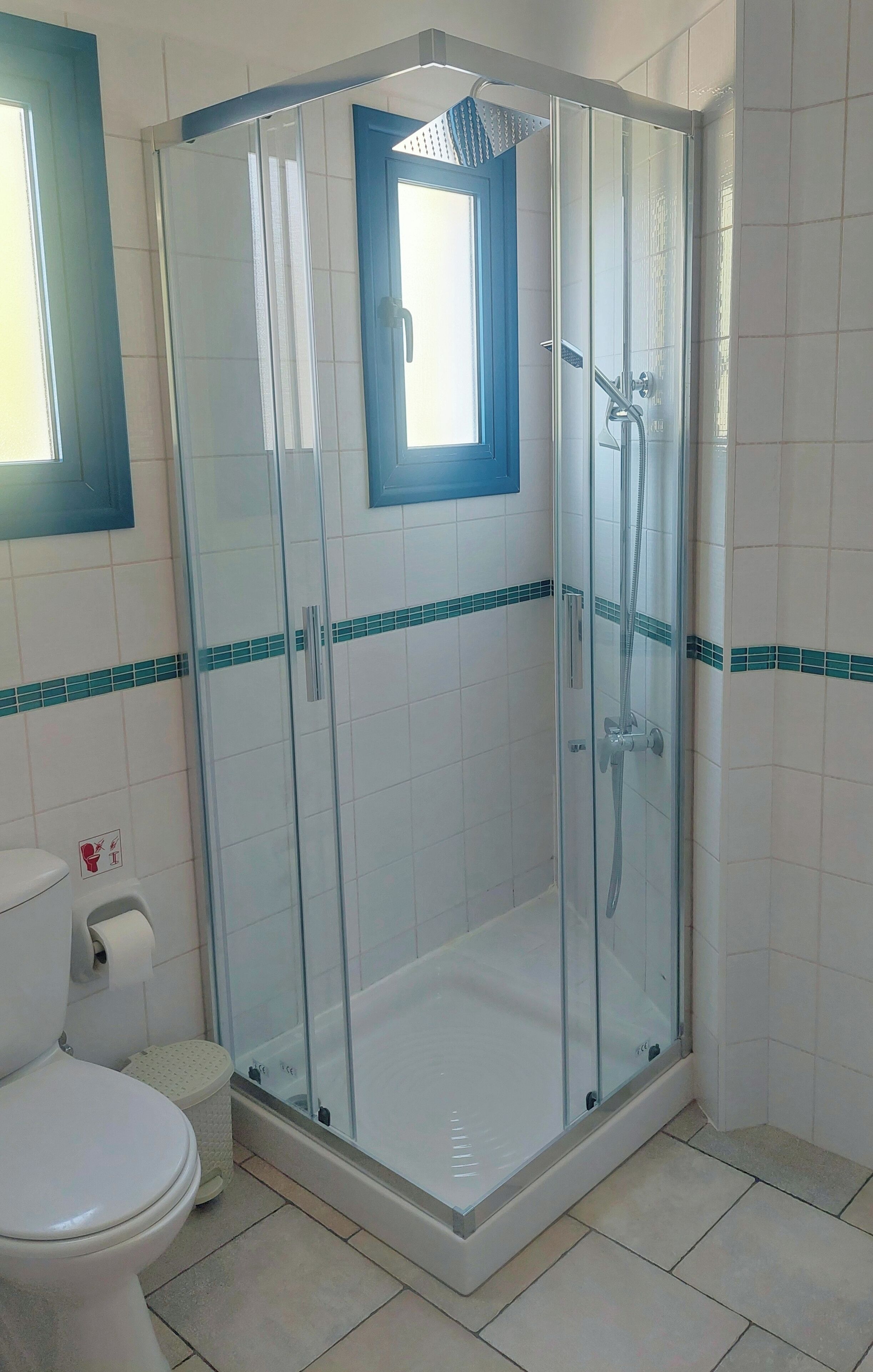 Box de ducha con mampara de vidrio, un inodoro y un cubo de basura en un baño con azulejos blancos y detalles verde mar.