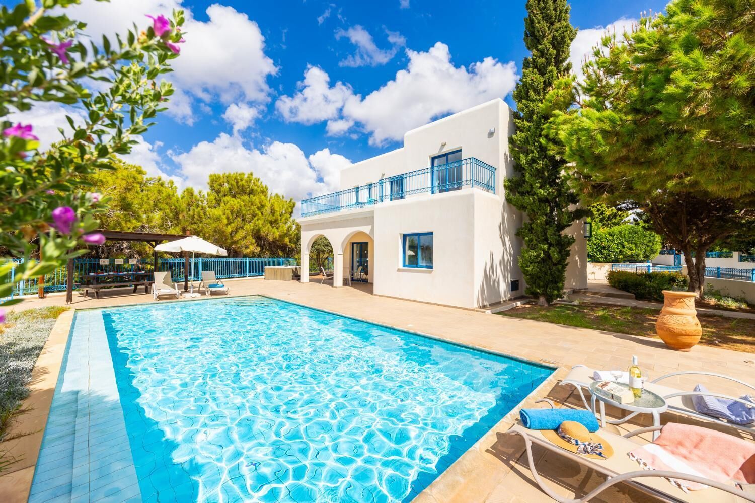A swimming pool with lounge chairs, a white building with blue accents, and surrounding greenery.
