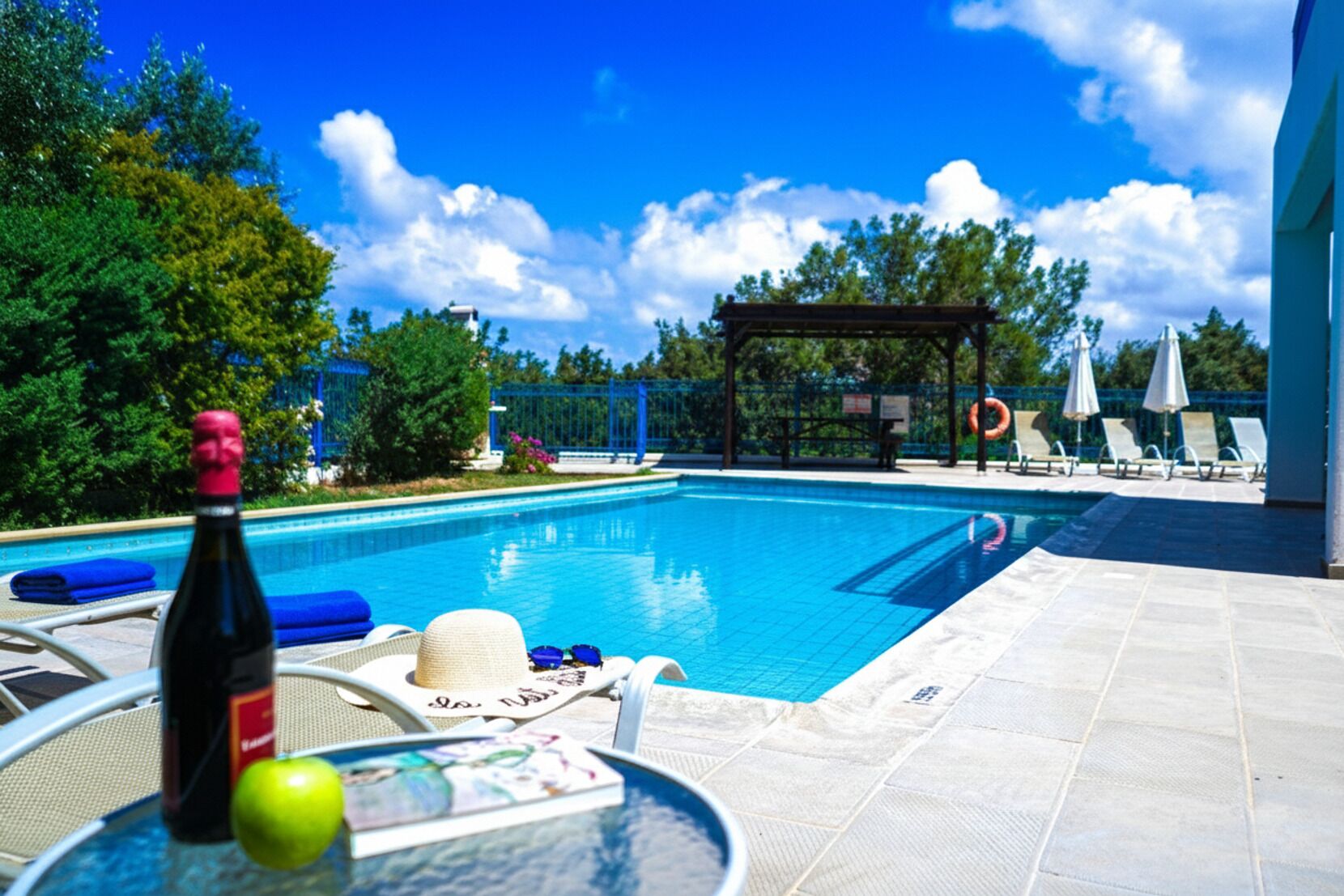 Une piscine avec des chaises longues et des parasols, une pergola et une bouteille de vin sur une table.