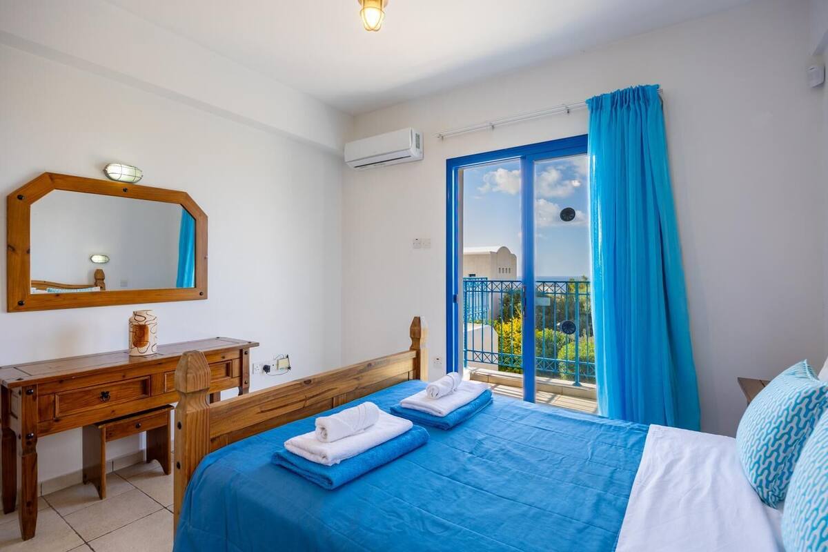 Een slaapkamer met een bed, een houten bureau, een spiegel en een balkon met blauwe gordijnen.