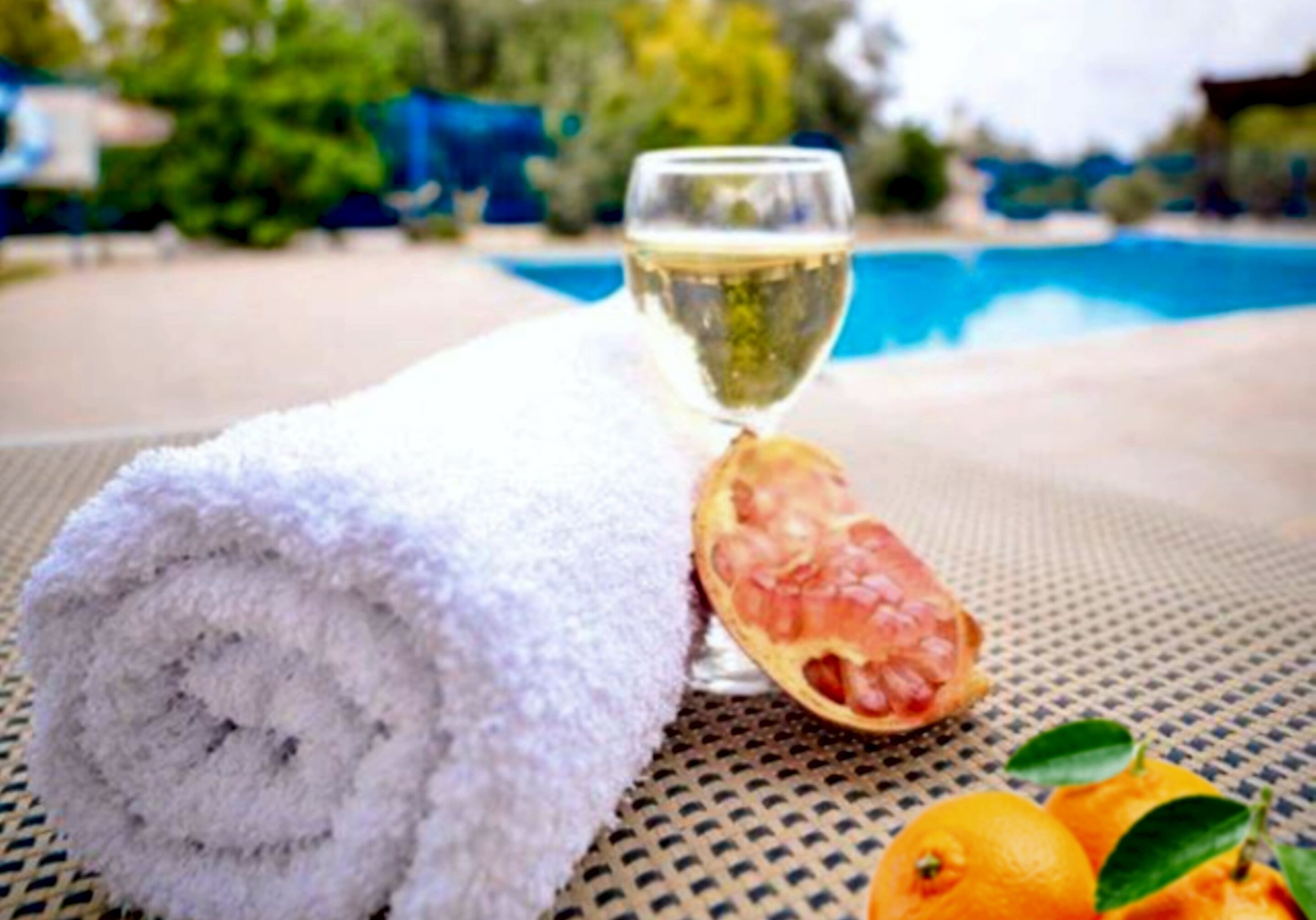 A towel, a glass of white wine, and a pomegranate on a table near a pool.