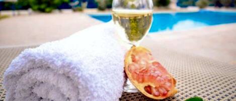 A towel, a glass of white wine, and a pomegranate on a table near a pool.