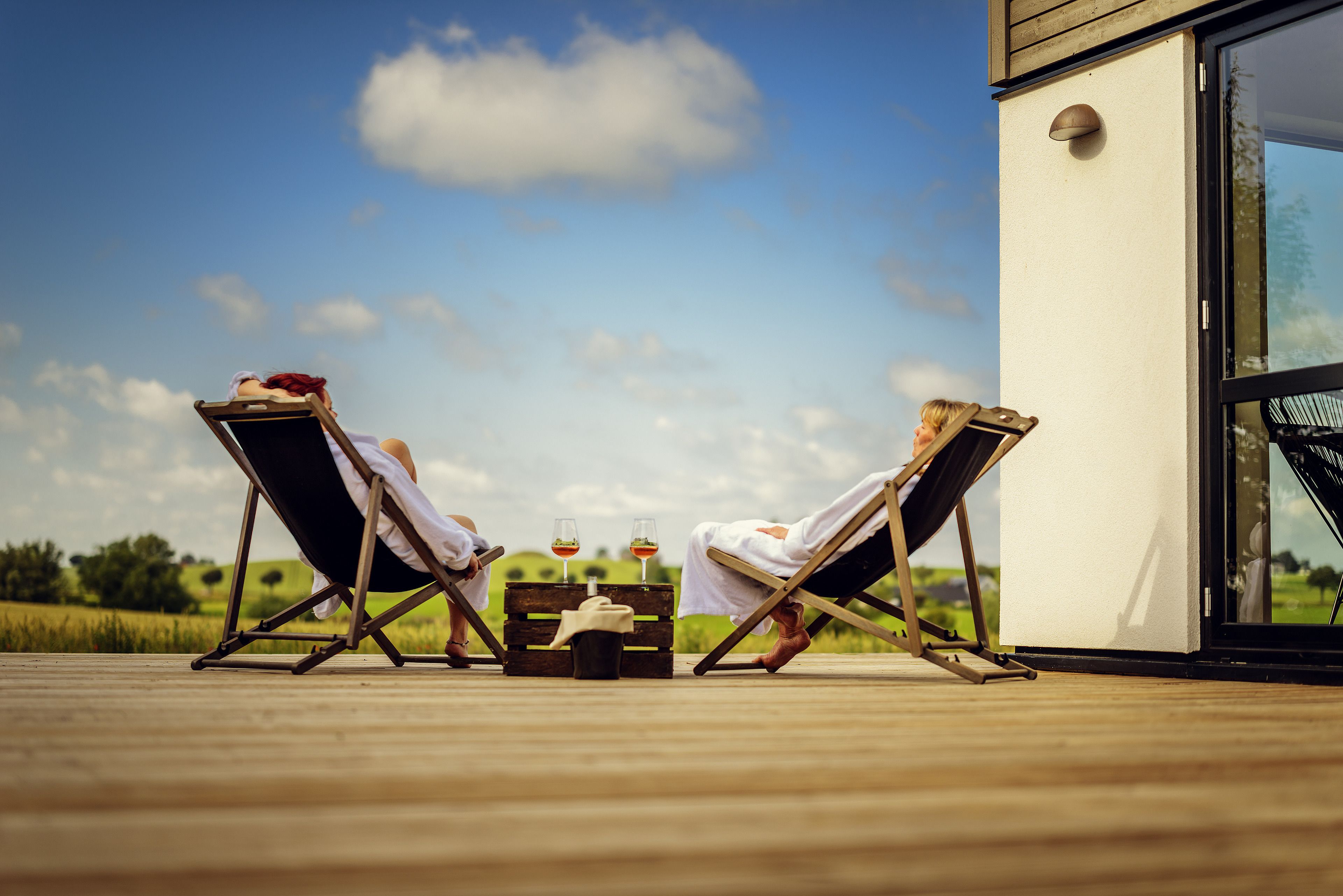 Two lounge chairs on a wooden deck with a table between them, a bottle, and two glasses of wine.