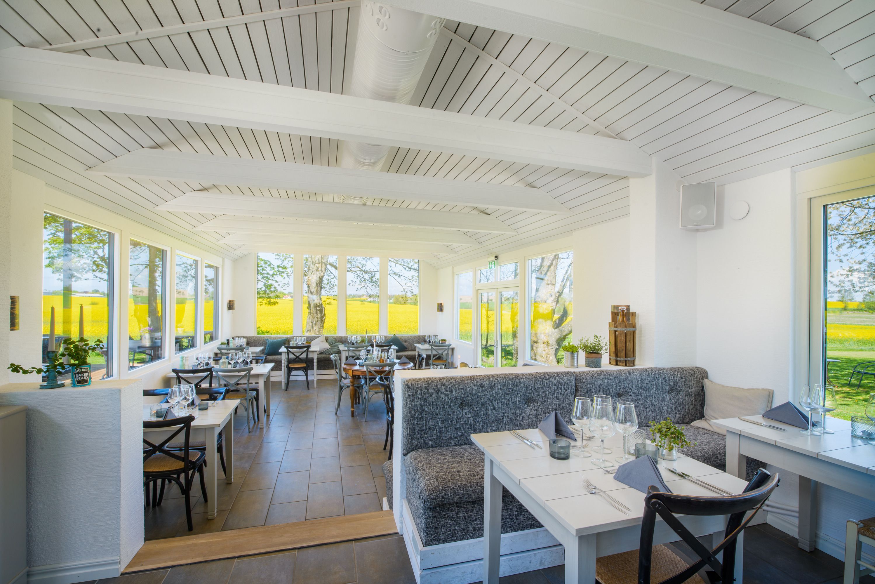 A bright, open-plan dining area with high ceilings and large windows. Tables are set for dining, and there are seating areas with cushions.