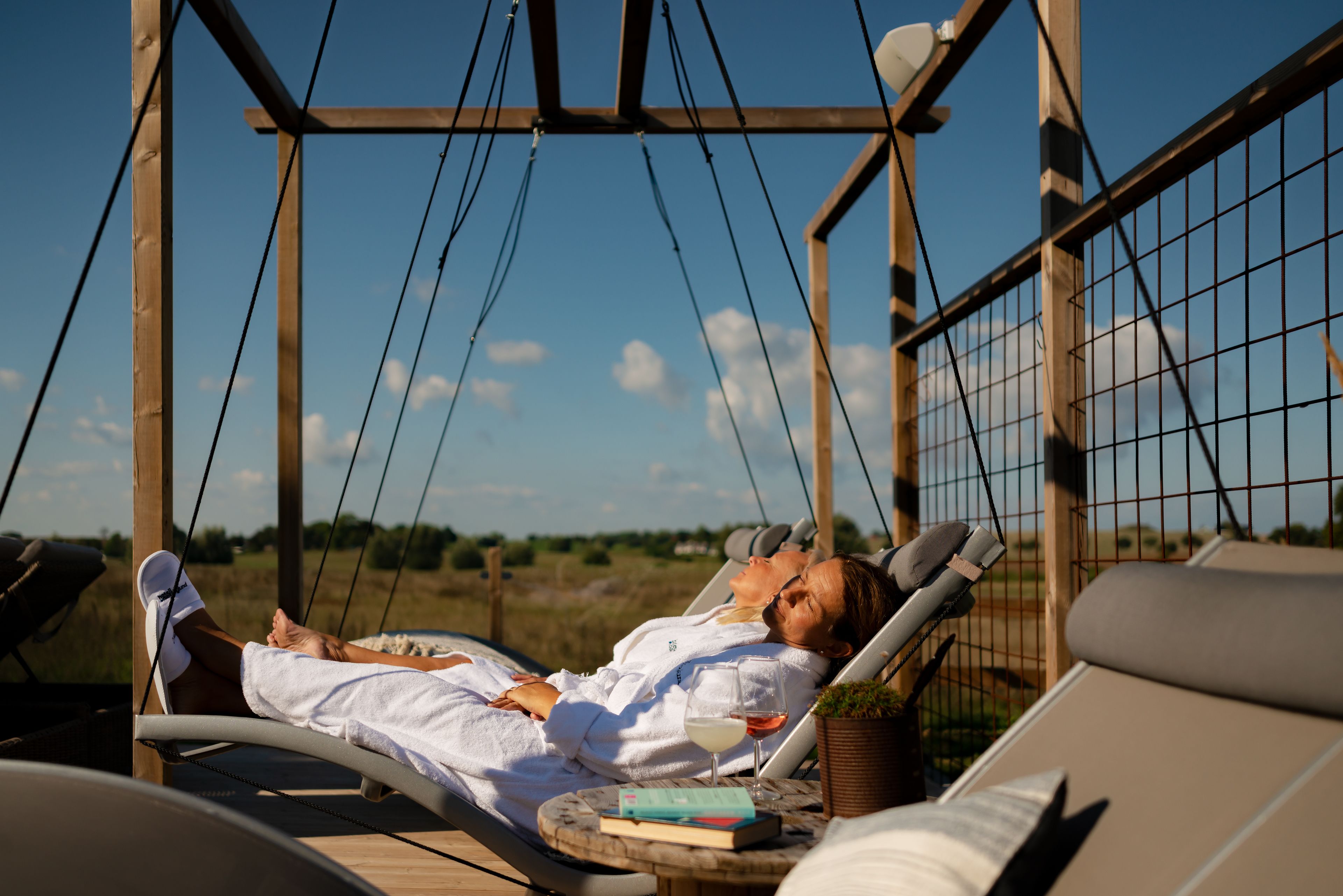 A couple relaxing in a hammock chair with a book and glass of wine, surrounded by wooden structures and a clear sky.