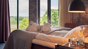 Une chambre d’hôtel avec une grande fenêtre, un lit avec des serviettes soigneusement pliées, une table de chevet avec une bouteille et des verres, et une lampe suspendue.