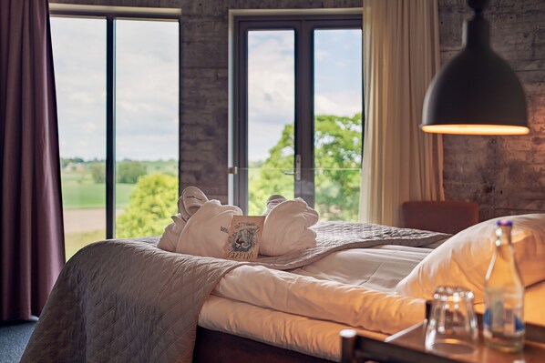 A hotel room with a large window, a bed with neatly folded towels, a bedside table with a bottle and glasses, and a hanging lamp. - Hotell Mossbylund (Skivarp)