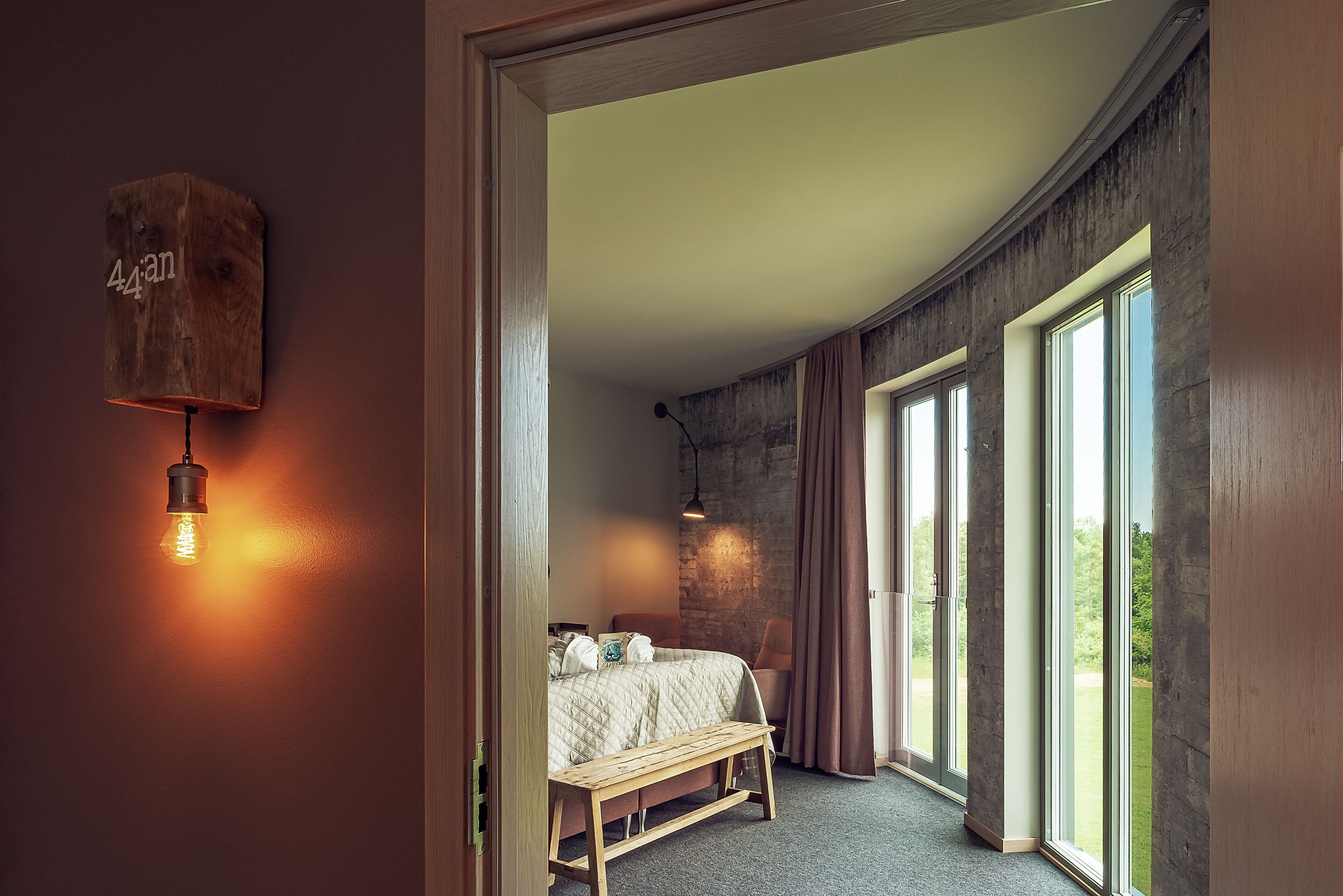 Un dormitorio con un ventanal grande, un banco de madera, una cama con colcha y una lámpara fijada a la pared.