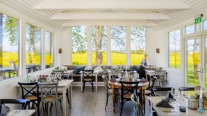 Un espace extérieur abrité, aménagé pour les repas, avec des tables et des chaises, de grandes fenêtres et une vue sur un champ.
