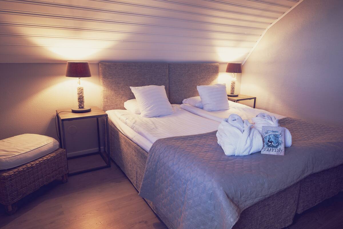 A hotel room with a large bed, two bedside tables, a small bench, and a ceiling with a sloped design.