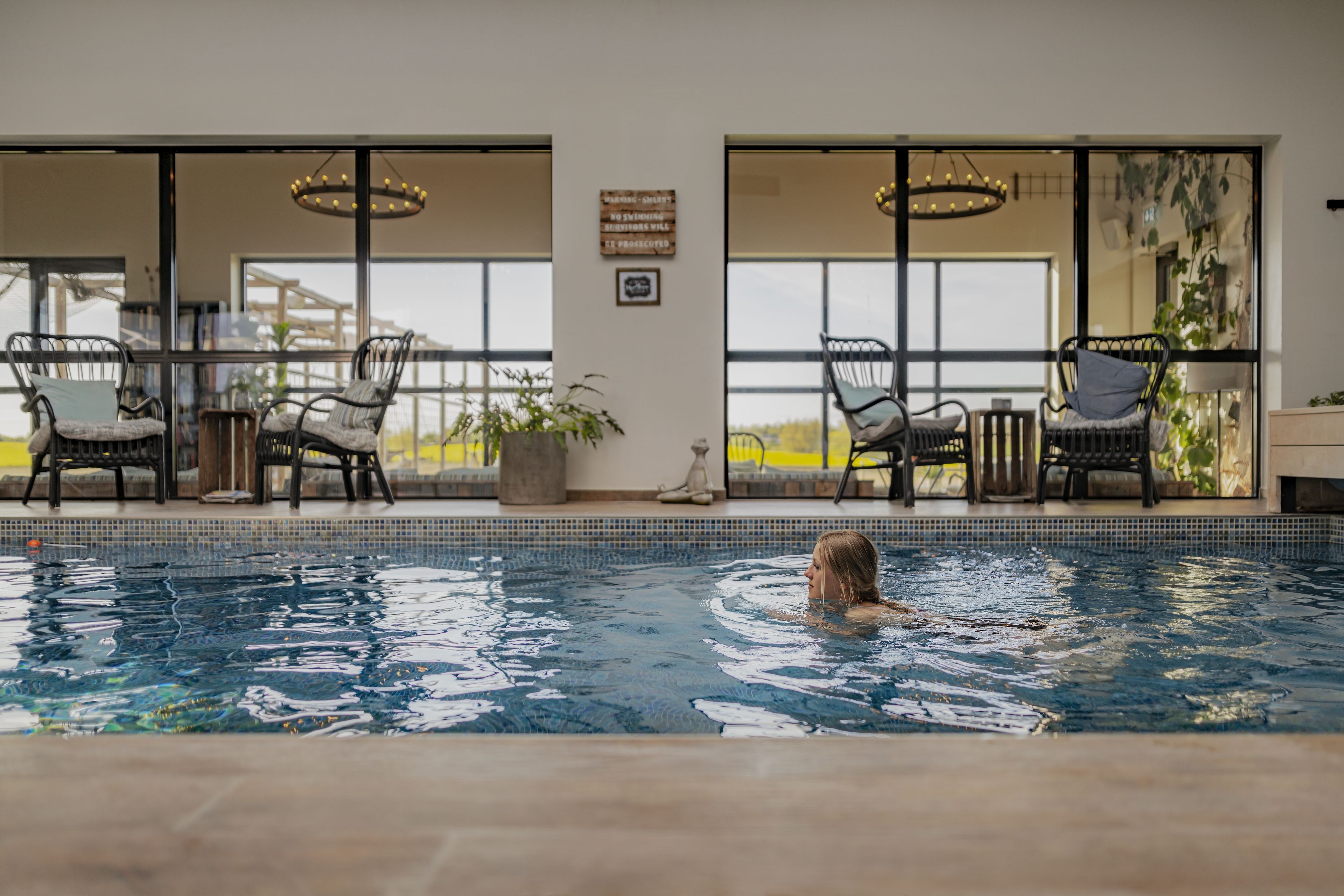 Una piscina interior con una persona nadando, rodeada de sillones de mimbre y una mesa, con grandes ventanales y vistas al exterior.