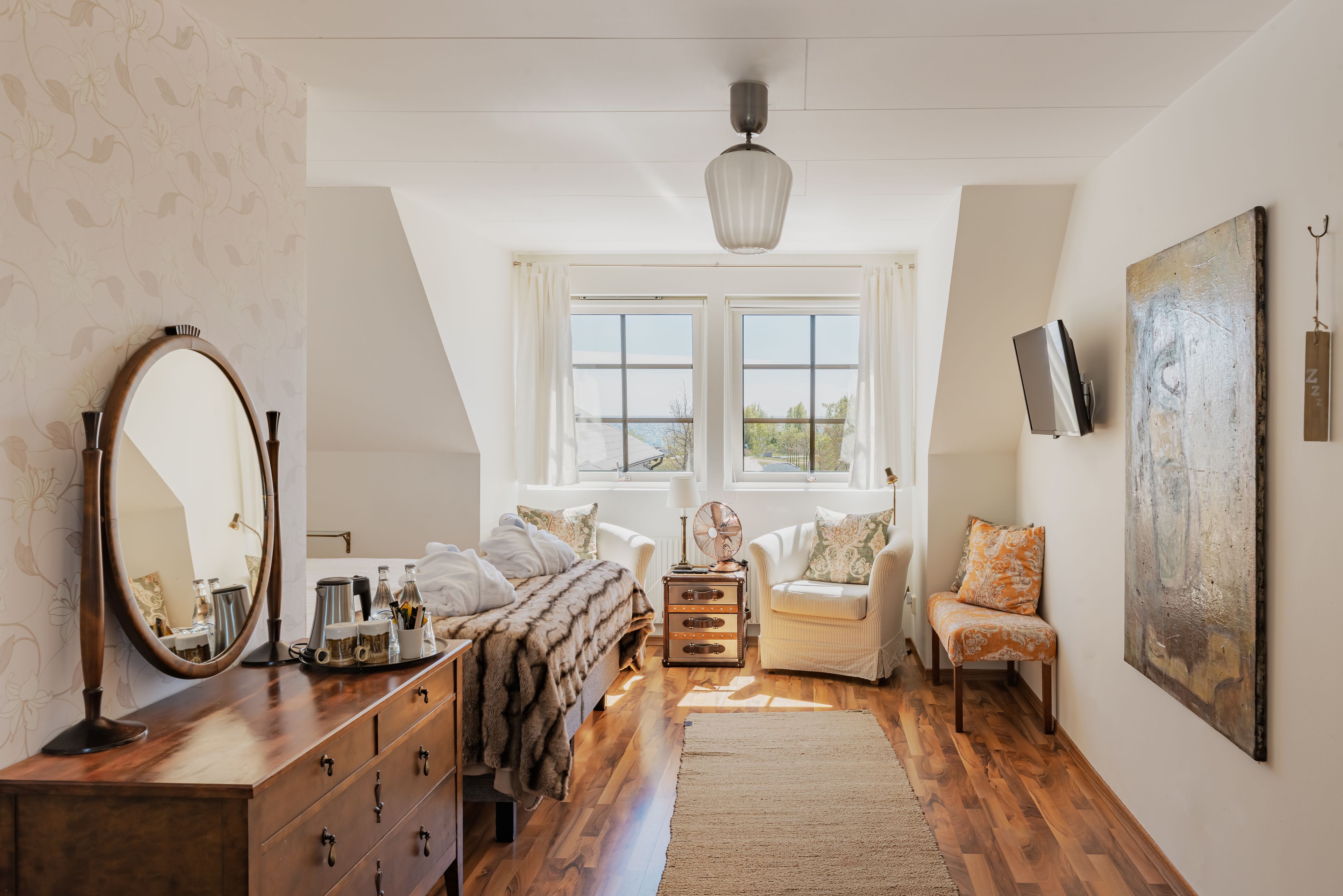 Quarto com cômoda de madeira, espelho grande, cama com manta de pele, duas cadeiras, janela com cortinas e um quadro na parede.
