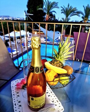 A bottle of champagne, a pineapple, a banana, and a basket of fruit on a table by a poolside.