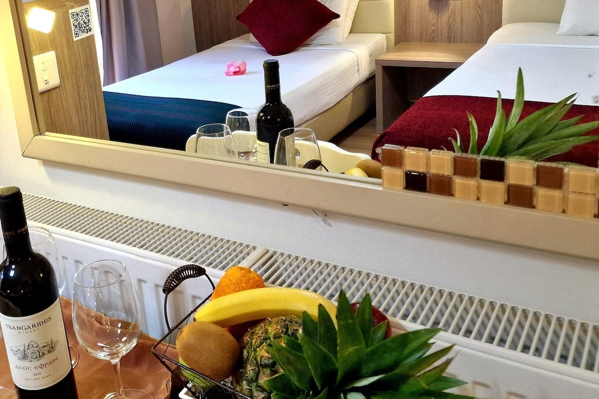 Et hotelværelse med to senge, et minikøleskab, et spejl der viser værelset, samt en frugtkurv med en flaske vin.