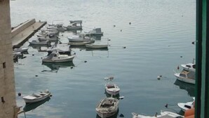 A harbor with multiple boats docked, a stone building with windows, and calm water. - XIV Century rooms (Kastela)