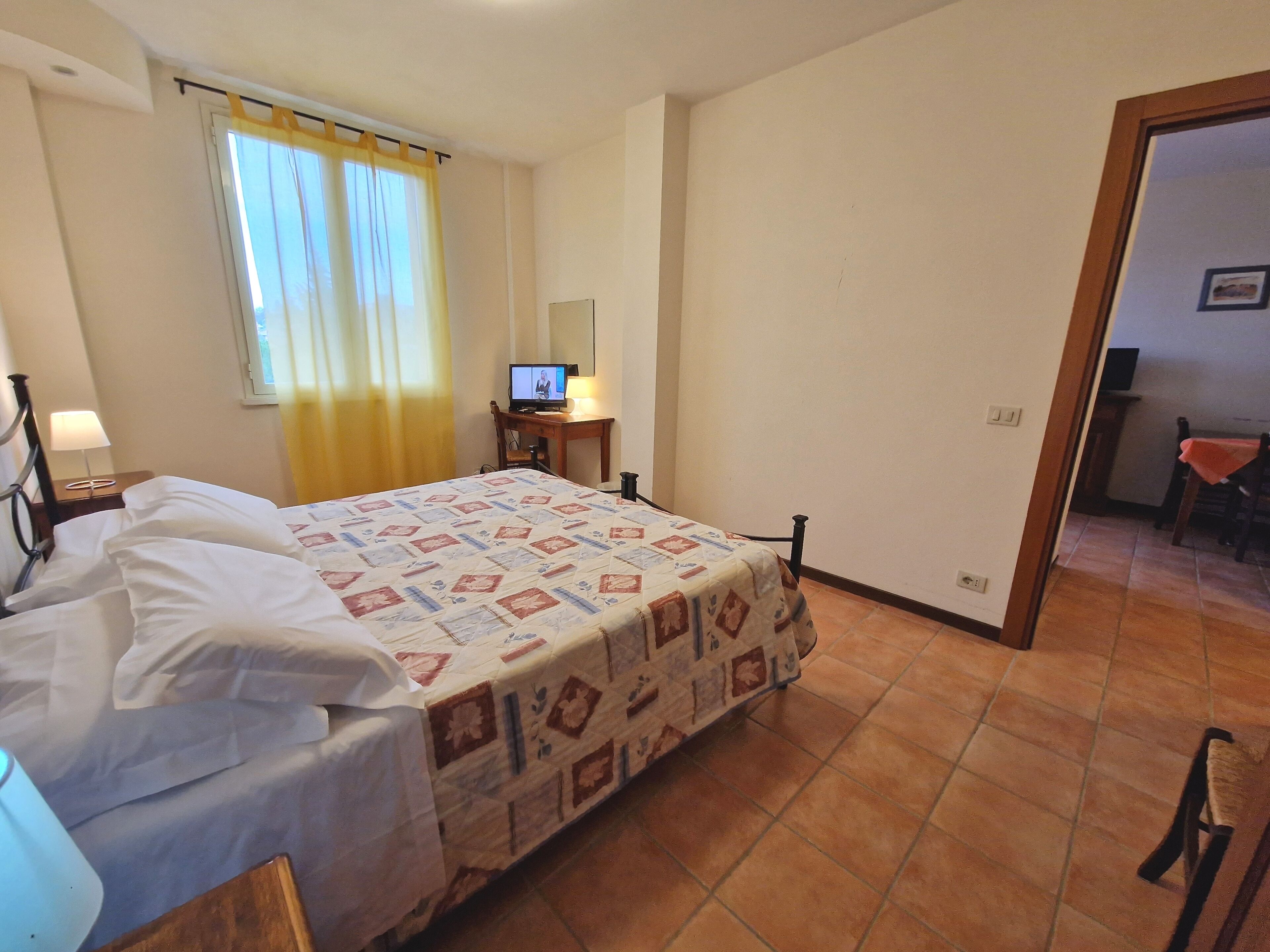 A hotel room with a bed, a desk with a computer, a chair, and a window with curtains.