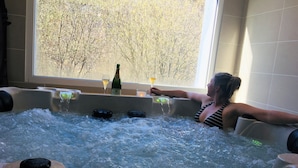 A woman relaxing in a hot tub with champagne and a glass of wine, with a view of trees outside.