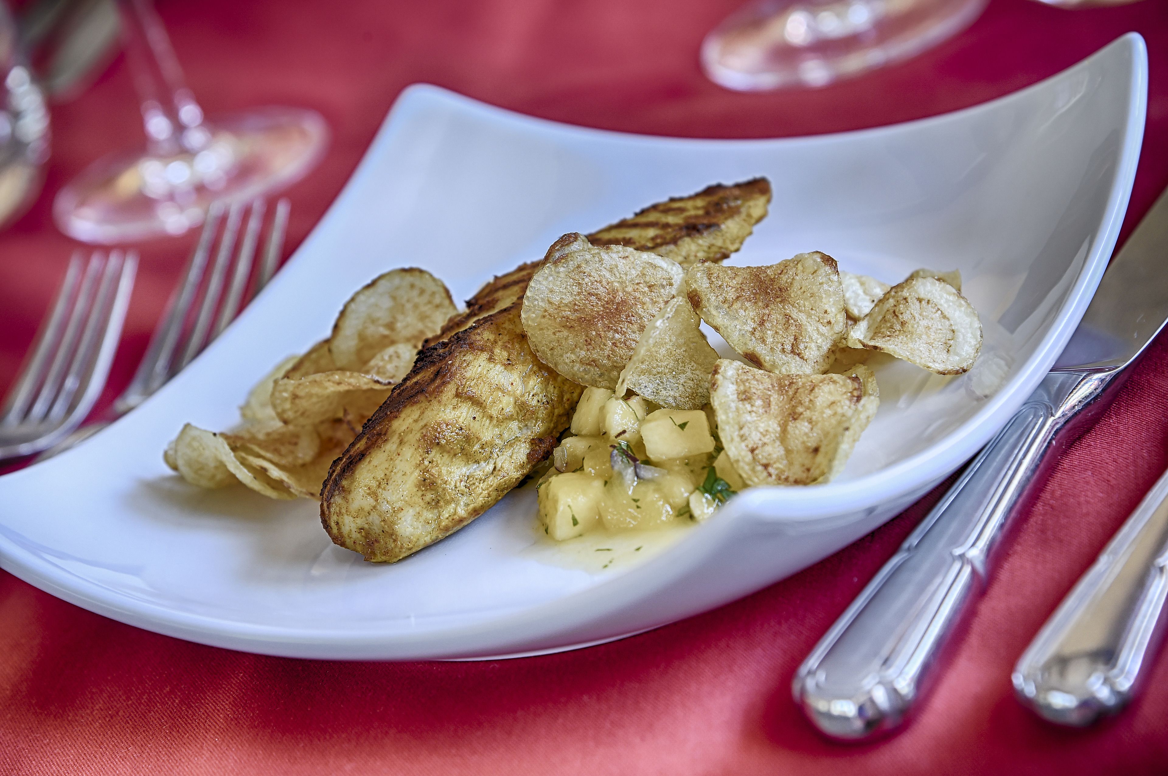 Une assiette blanche contenant des chips et un petit accompagnement, avec des couverts posés sur une nappe rouge.