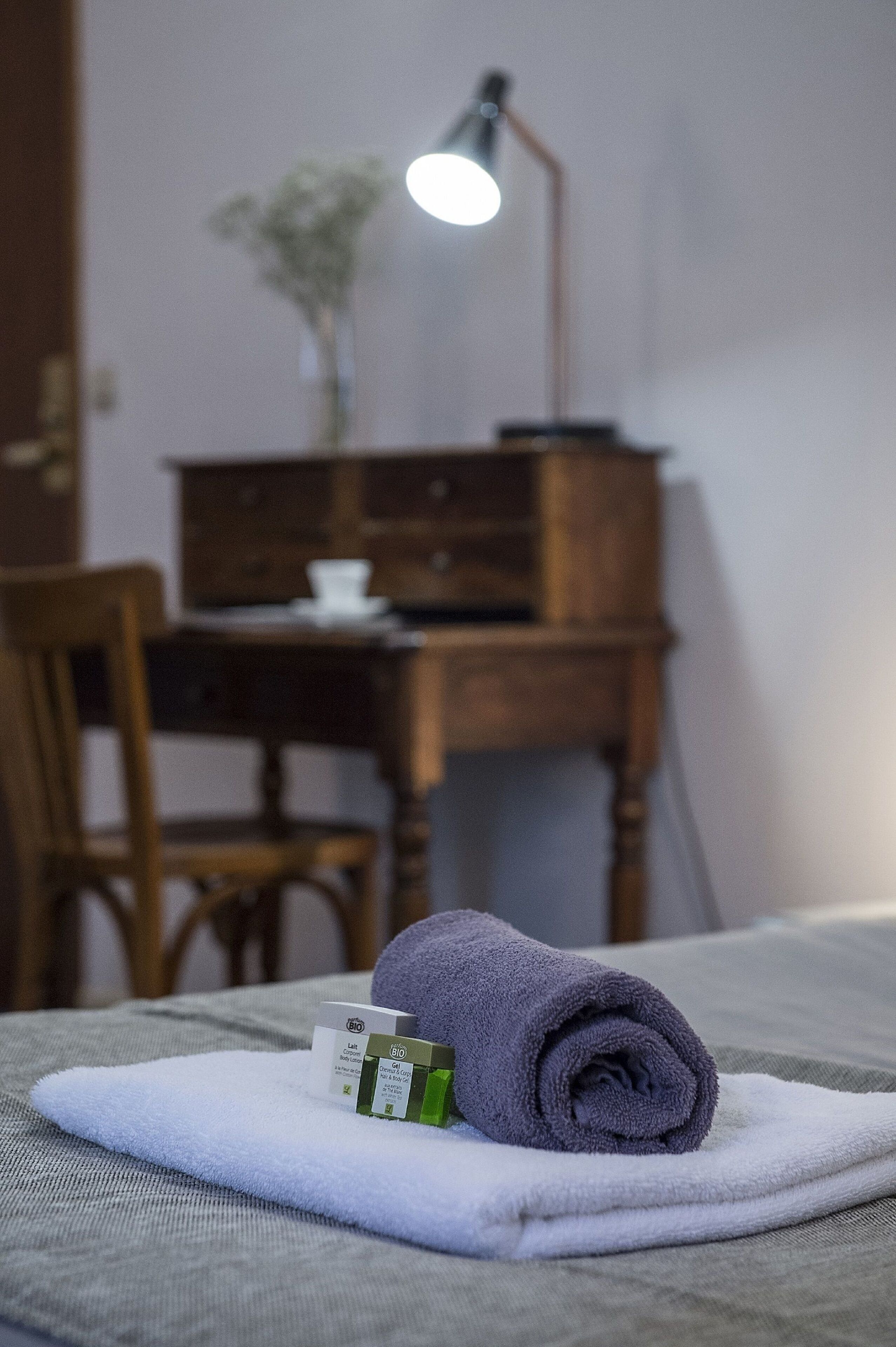 A neatly made bed with a rolled towel and a small box, a wooden desk with a lamp, and a chair in a room with a simple, clean design.