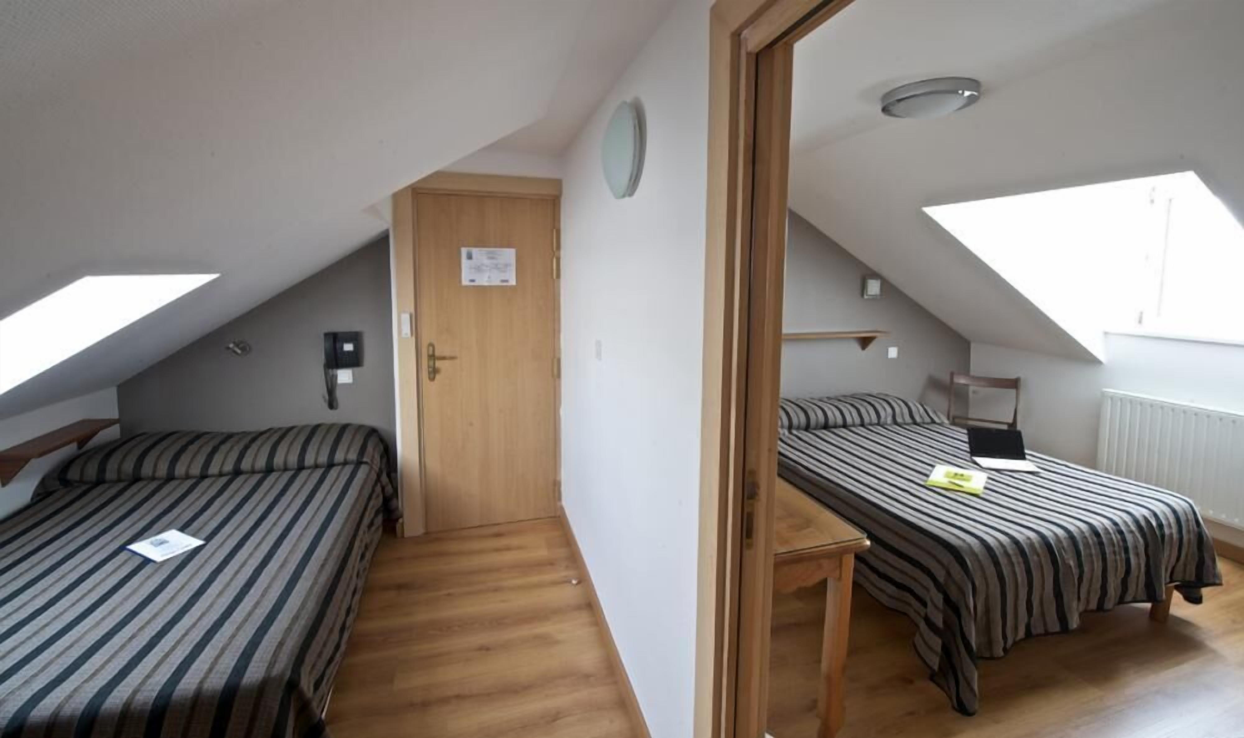 Une chambre de dortoir avec deux lits simples, une porte en bois, une petite table et un puits de lumière.