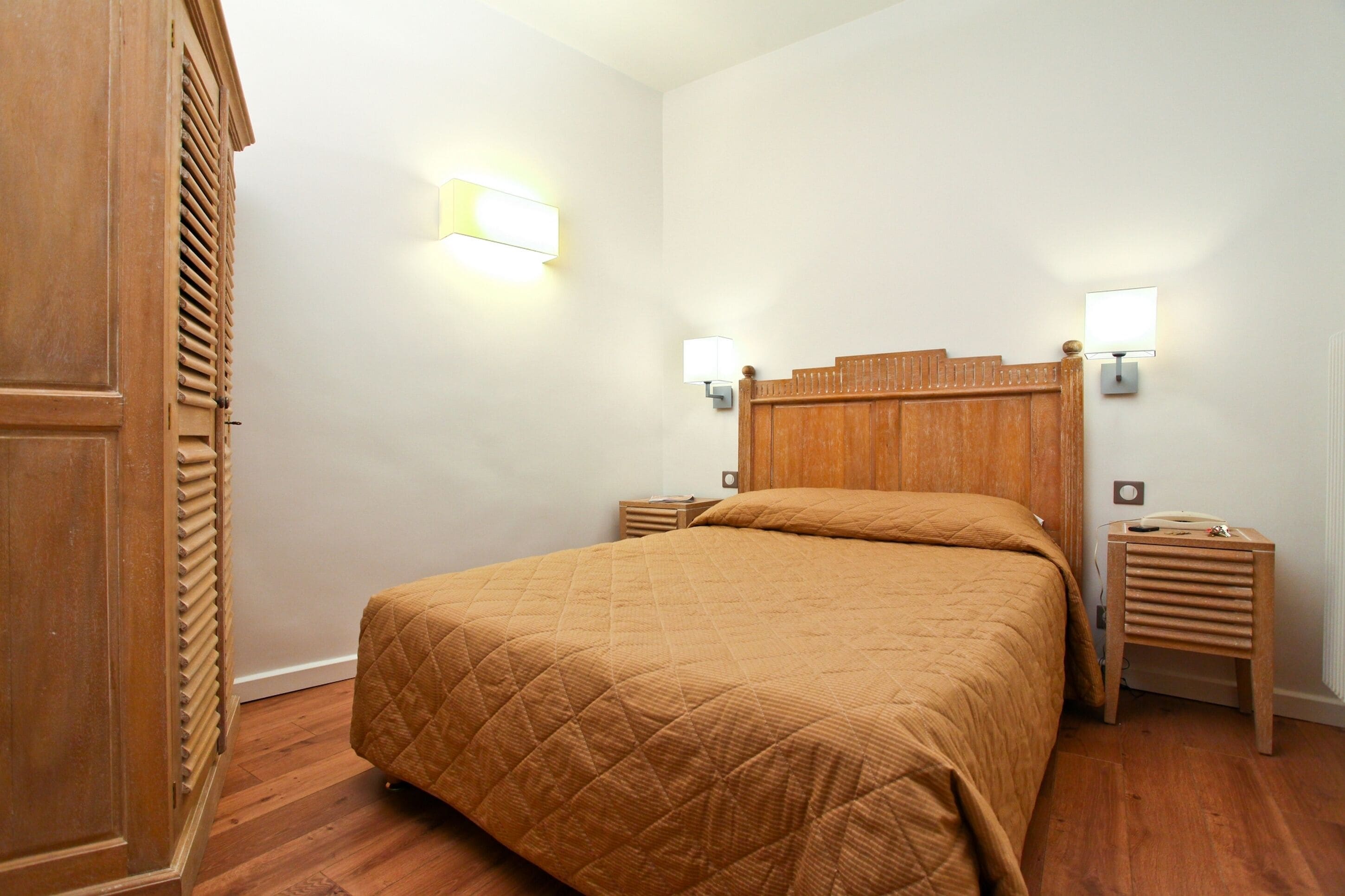 Un dormitorio con cabecera de madera, colcha acolchada, dos mesitas de noche con lámparas y suelo de madera.