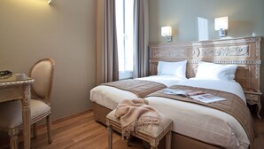 Quarto de hotel com uma cama grande, duas luminárias de cabeceira, um painel de madeira na cabeceira, uma cadeira e uma pequena mesa com uma luminária.