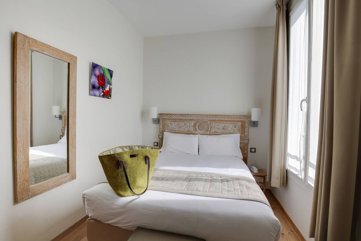 A neatly arranged hotel room with a large bed, a bedside table, a nightstand, a mirror, and a window with curtains.