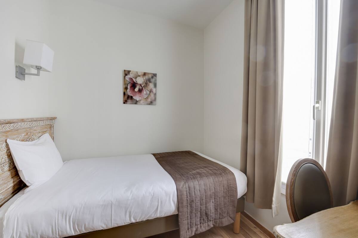 A neatly made bed with a white and brown bedspread, a wall-mounted light fixture, and a floral painting.