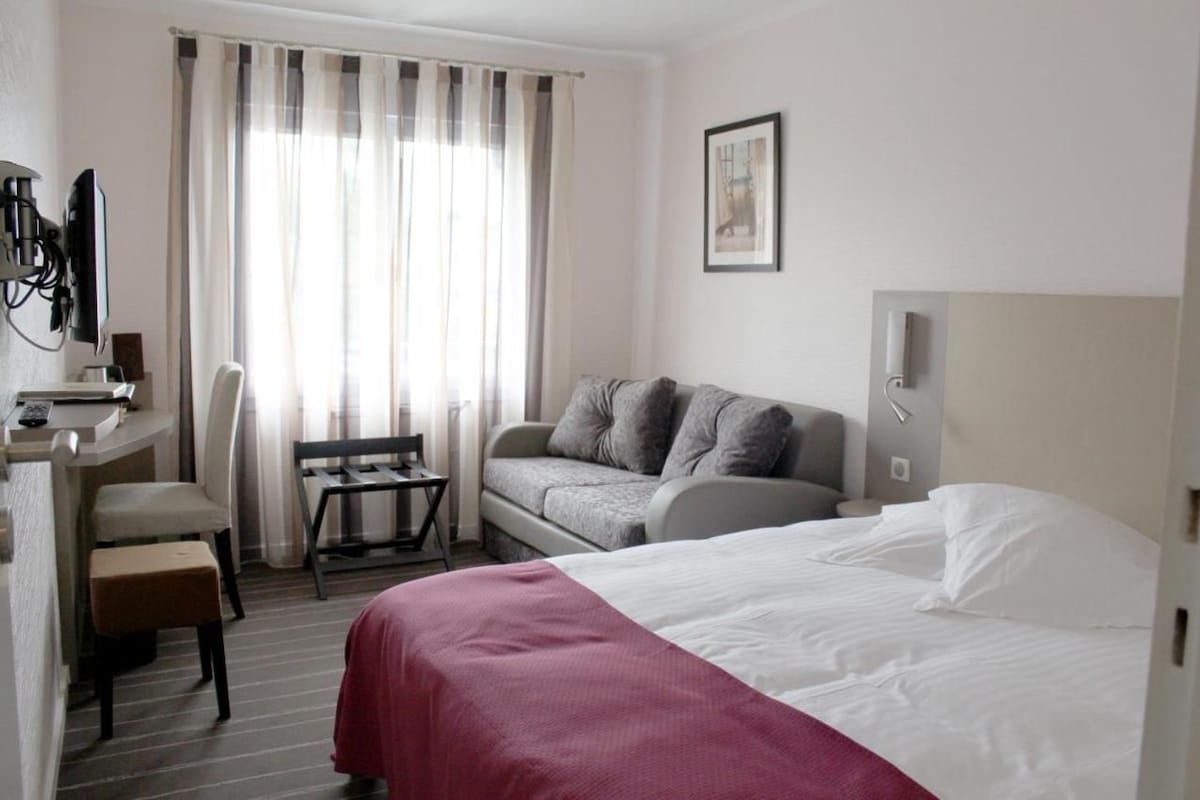 A hotel room with a bed, a sofa, a desk with a chair, a television, and a window with curtains.