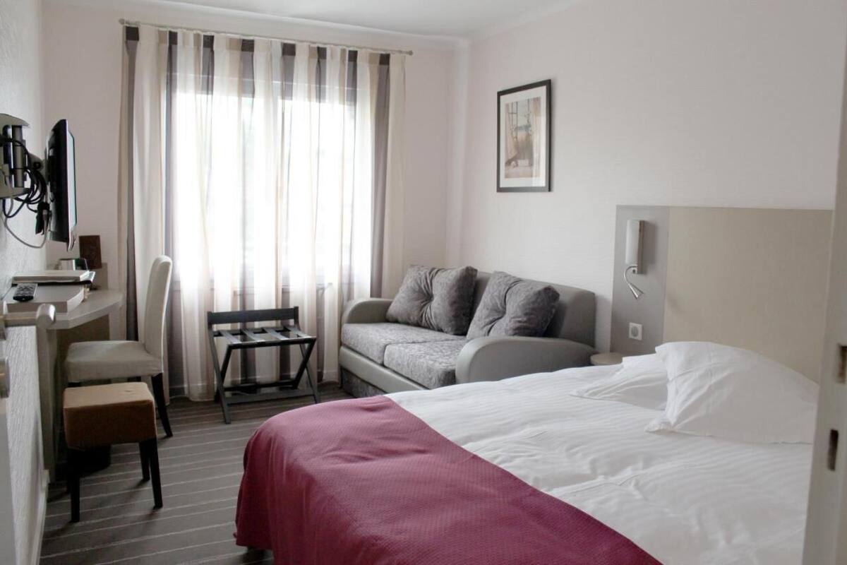 A hotel room with a bed, a sofa, a desk with a chair, a television, and a window with curtains.