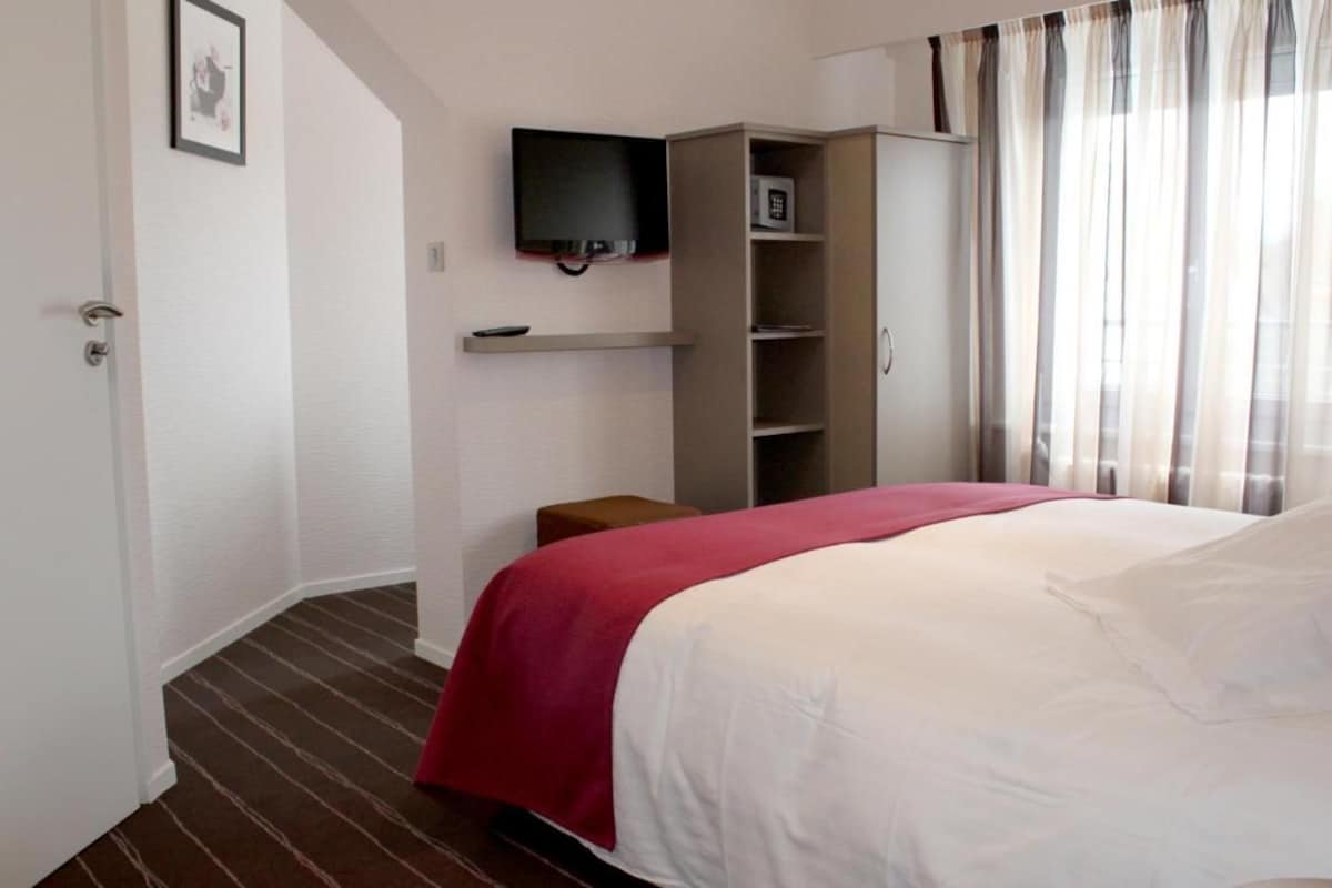 A hotel room with a bed, a television mounted on the wall, a wardrobe, and a small stool.
