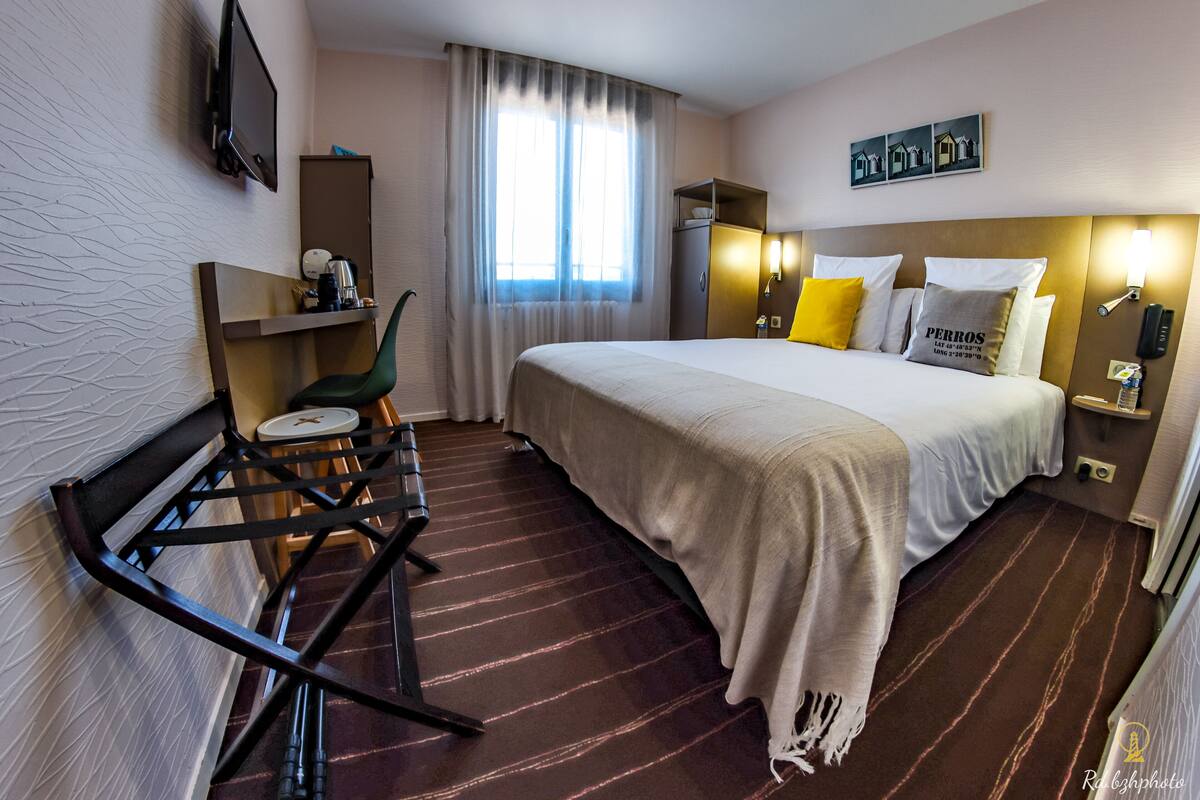 A hotel room with a bed, a desk with a chair, a television, and a window with curtains.