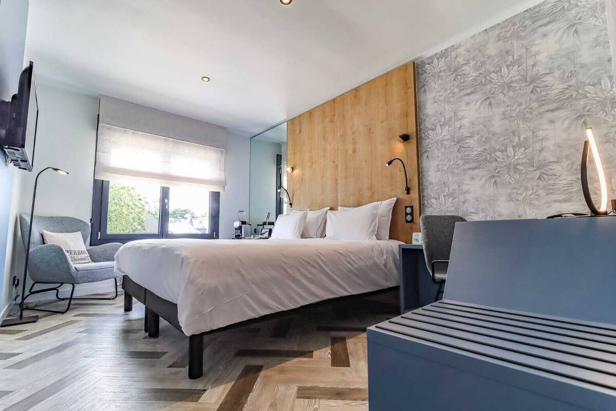 A modern hotel room with a large bed, a desk, a chair, and a view of greenery outside.