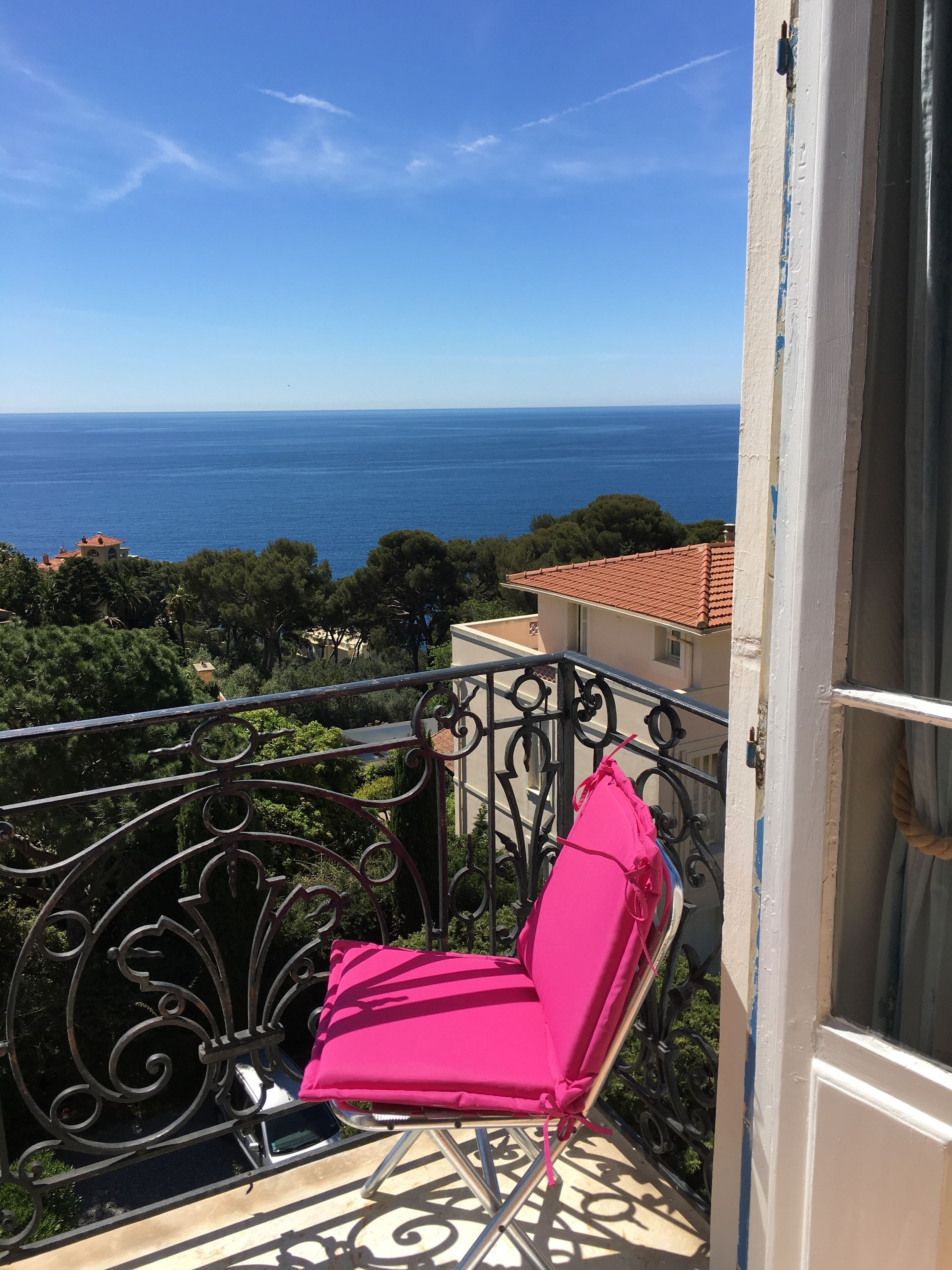Un balcon avec une chaise rose, une vue sur la mer et un bâtiment avec un toit recouvert de tuiles rouges.