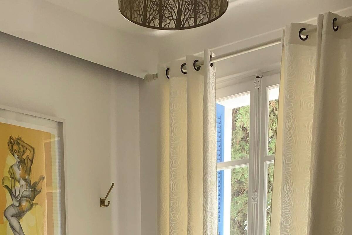 A hotel room with a bed, pillows, a nightstand, a painting, and a window with curtains.
