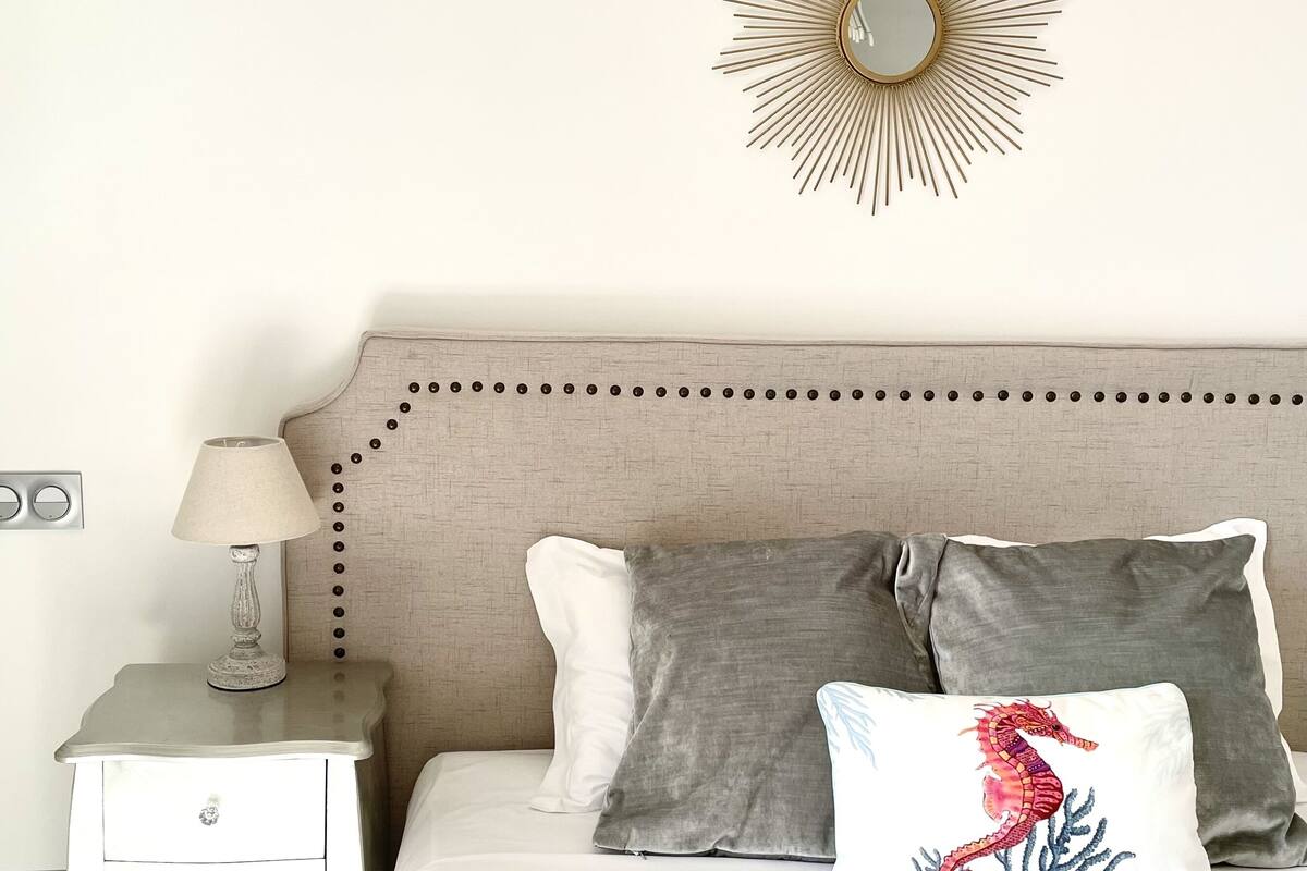 A neatly arranged bedroom with a bed, pillows, a nightstand, a lamp, and a wall-mounted mirror.