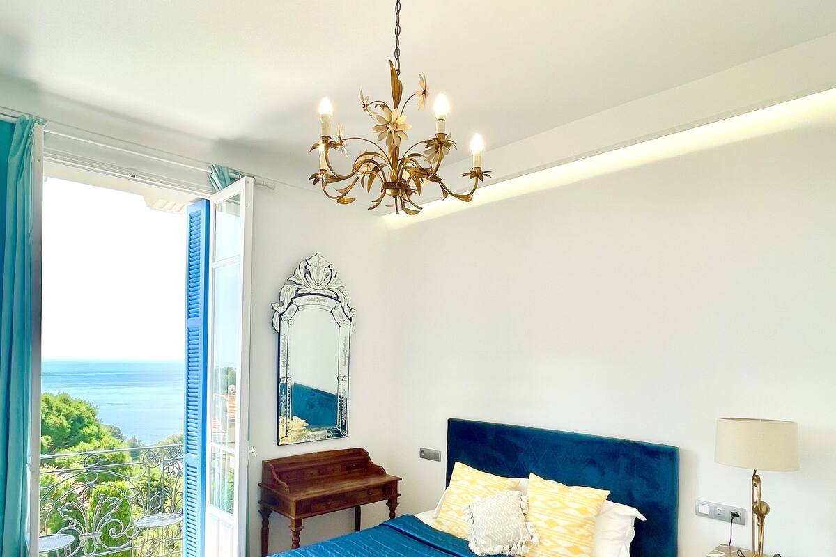 A bedroom with a large bed, a chandelier, a nightstand, and a view of the sea.