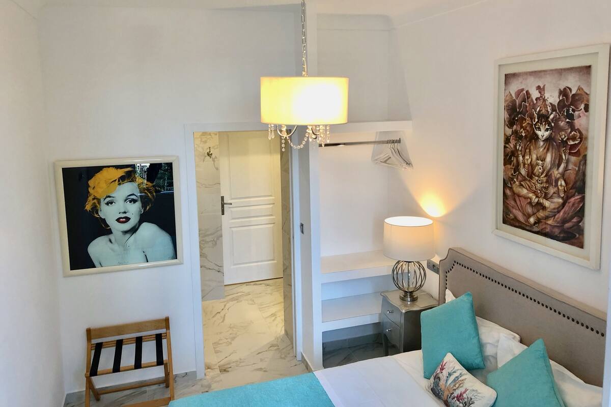 A hotel room with a bed, a bench, a nightstand with a lamp, a chandelier, and two framed artworks on the walls.