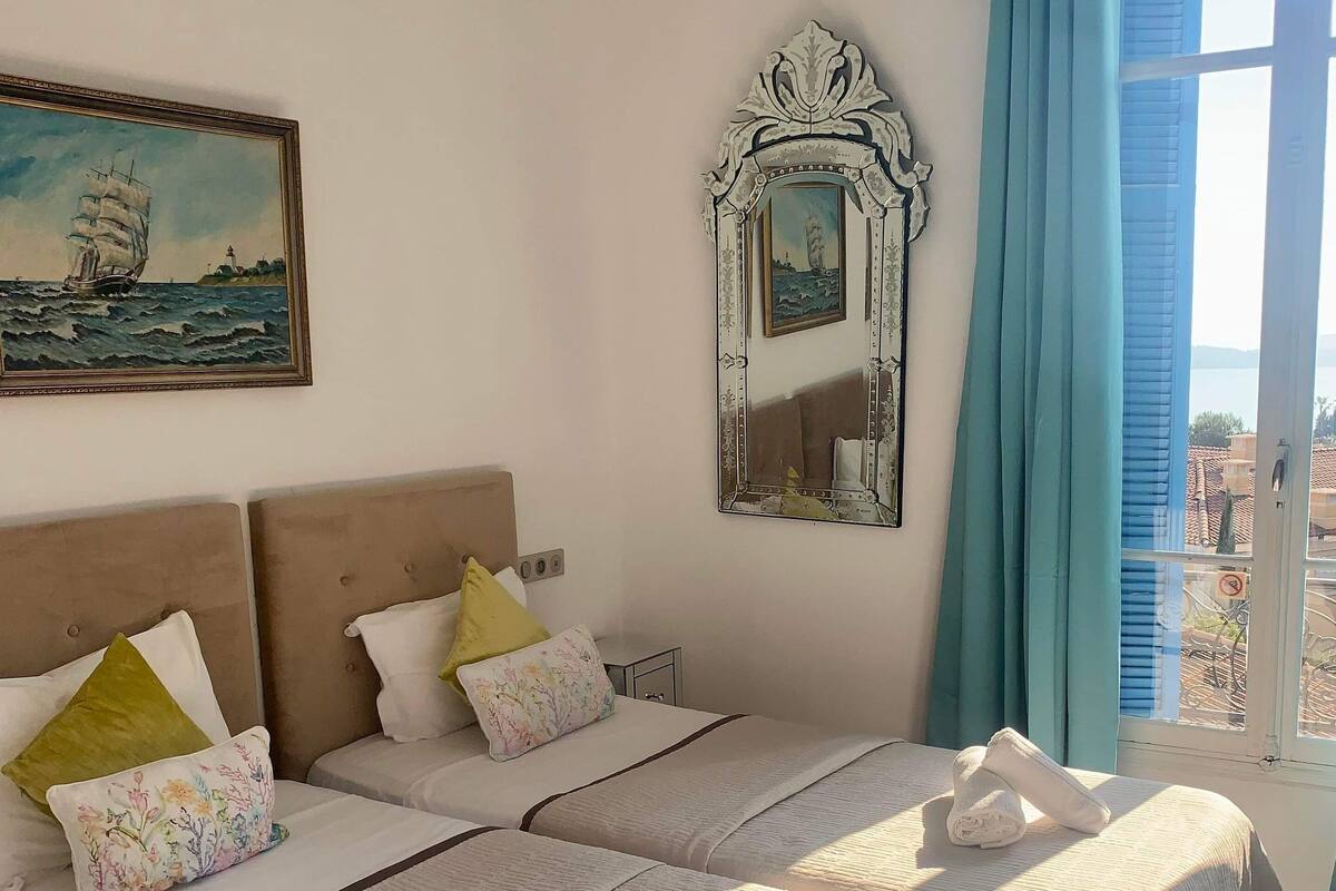 A hotel room with two beds, a painting, a mirror, and a view of the sea.
